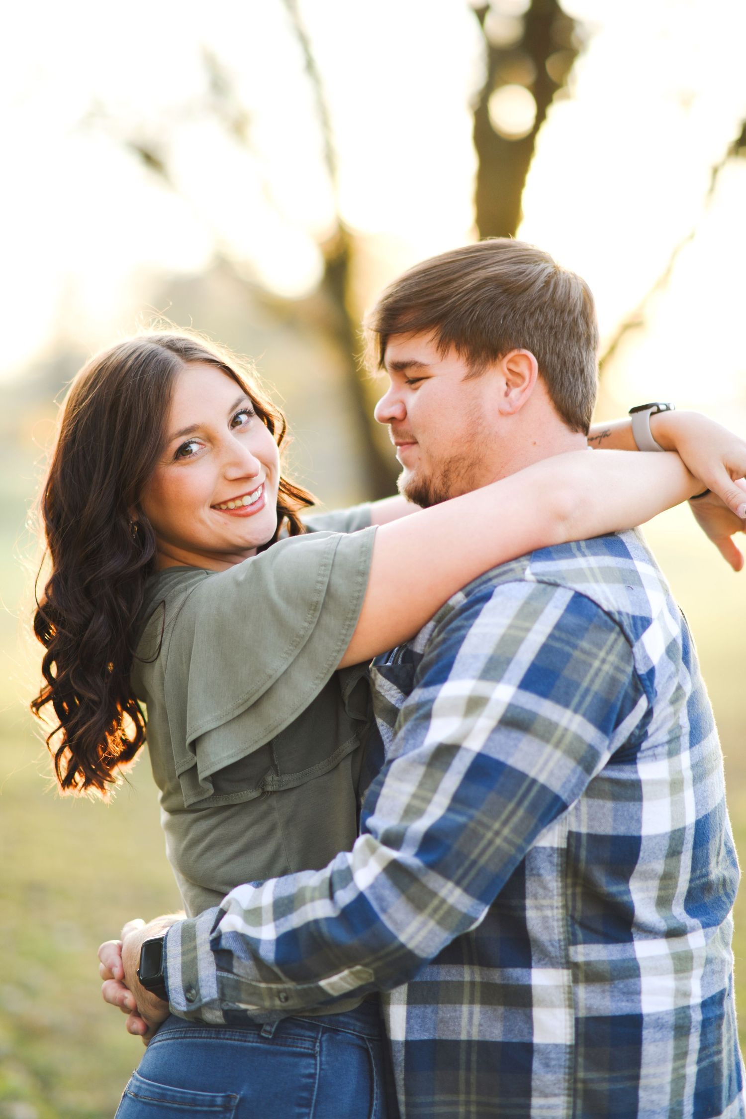Woman smiling at camera while embracing man outdoors in warm golden hour sunlight in a park.