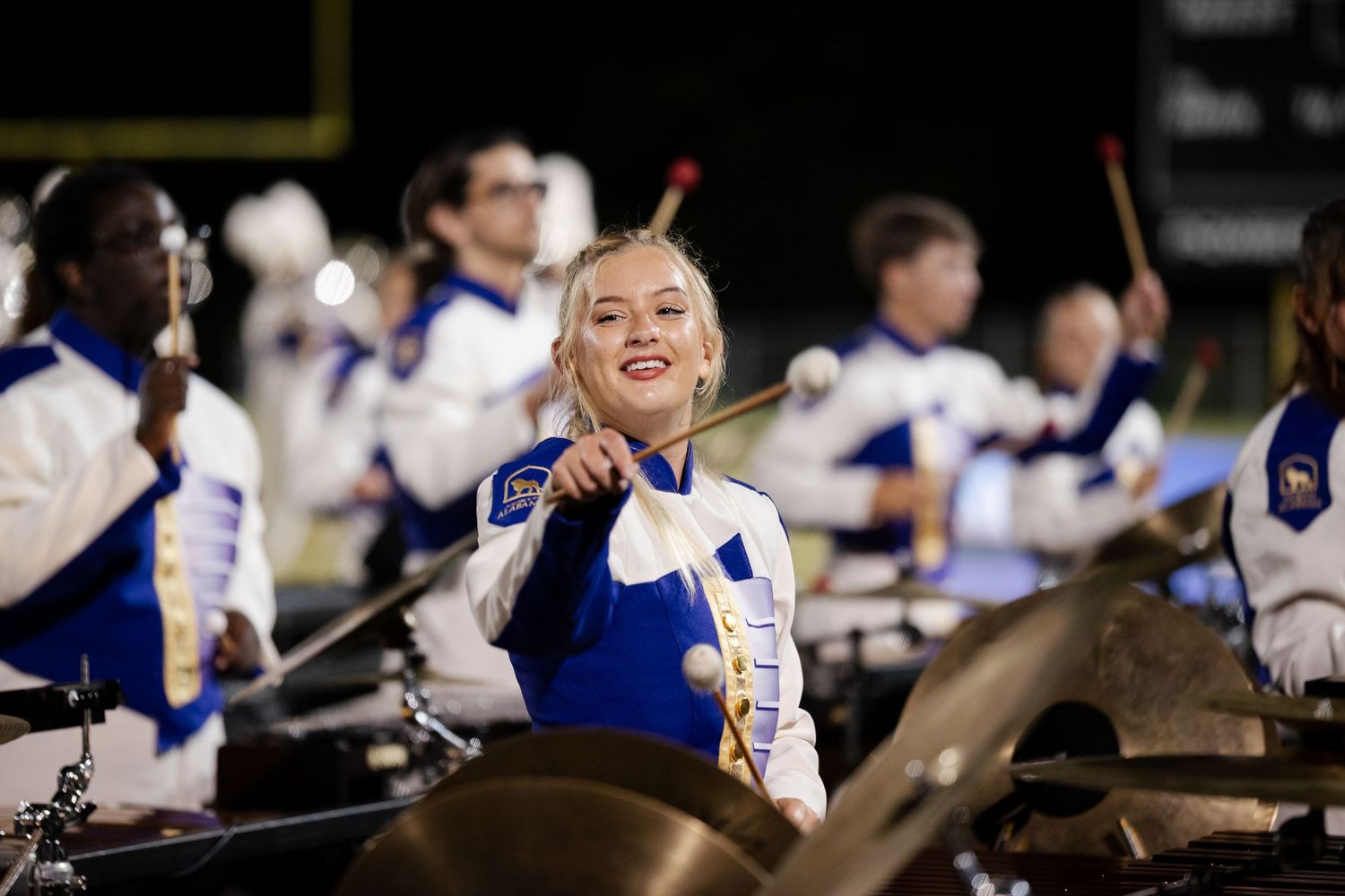 A smiling female percussionist plays timpani drums during a nighttime marching band performance.