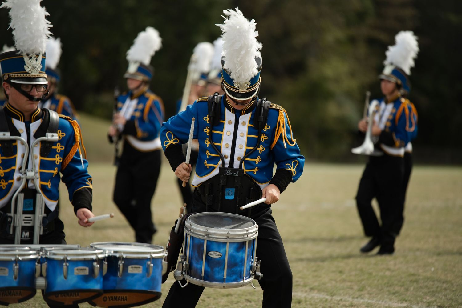 Marching band drummer in blue and gold uniform performs with snare drums on a field.