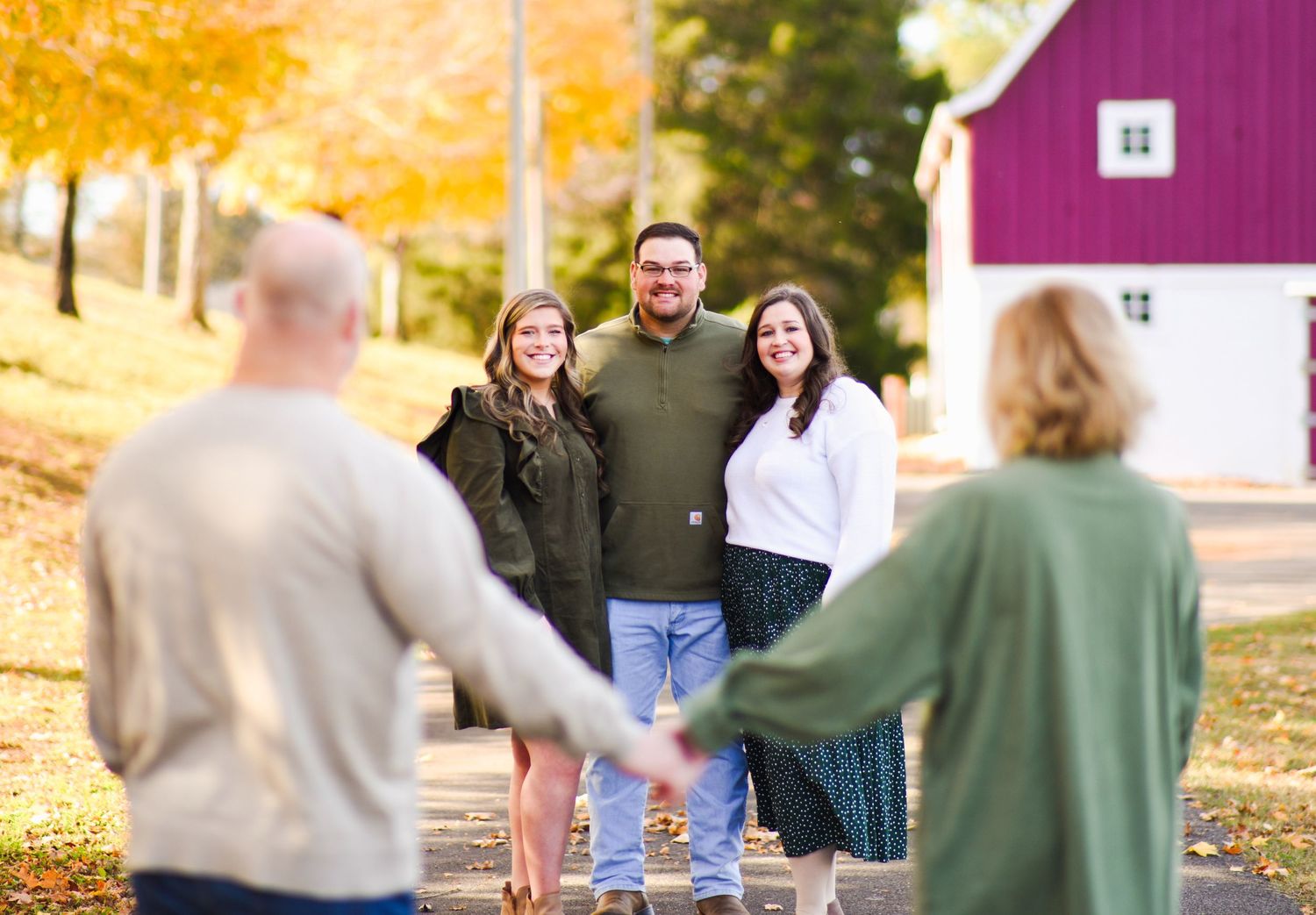 Group of four adults smiles together on a tree-lined path near a red barn in autumn.