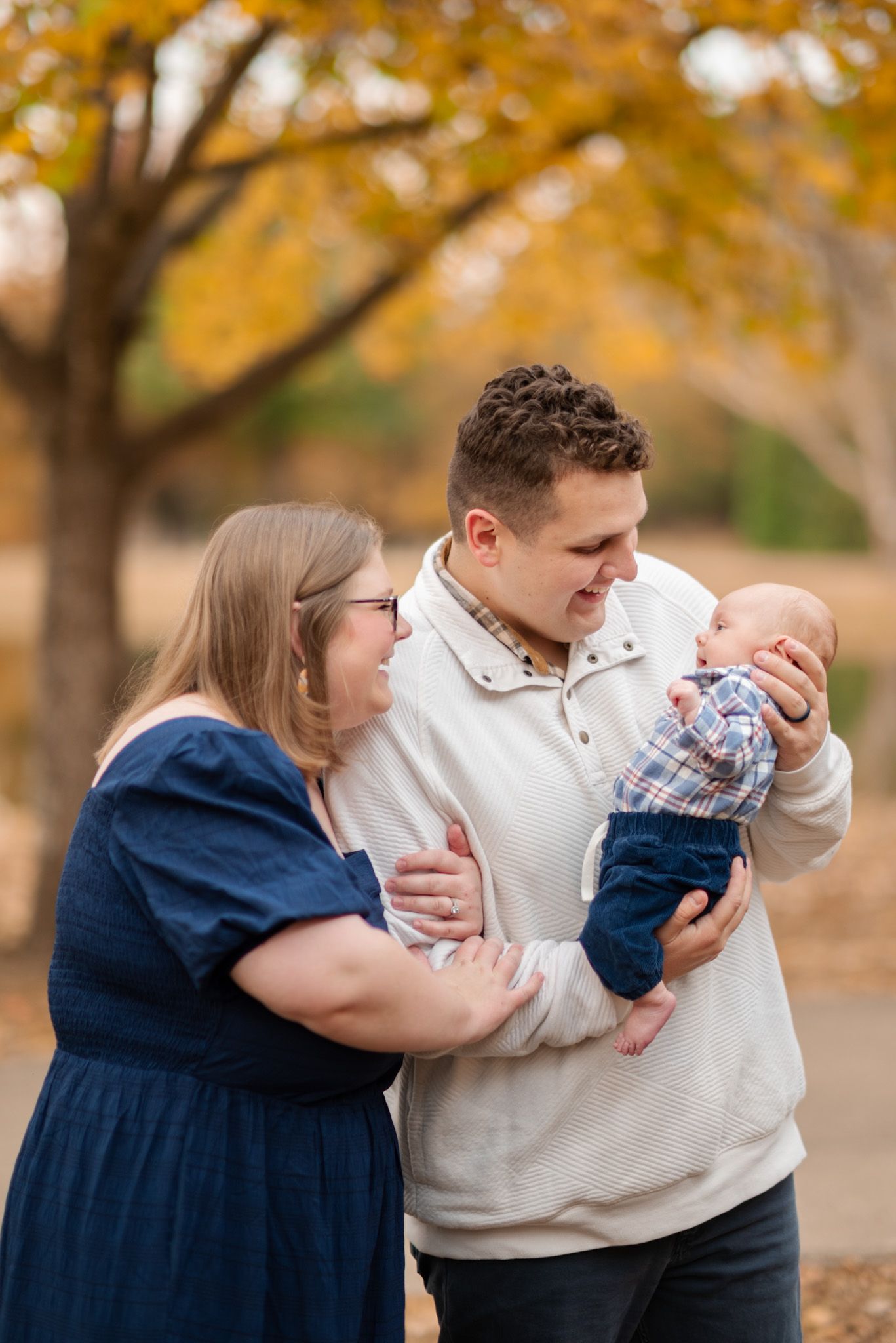 Young couple lovingly gazing at newborn baby held by father during fall outdoor family portrait.