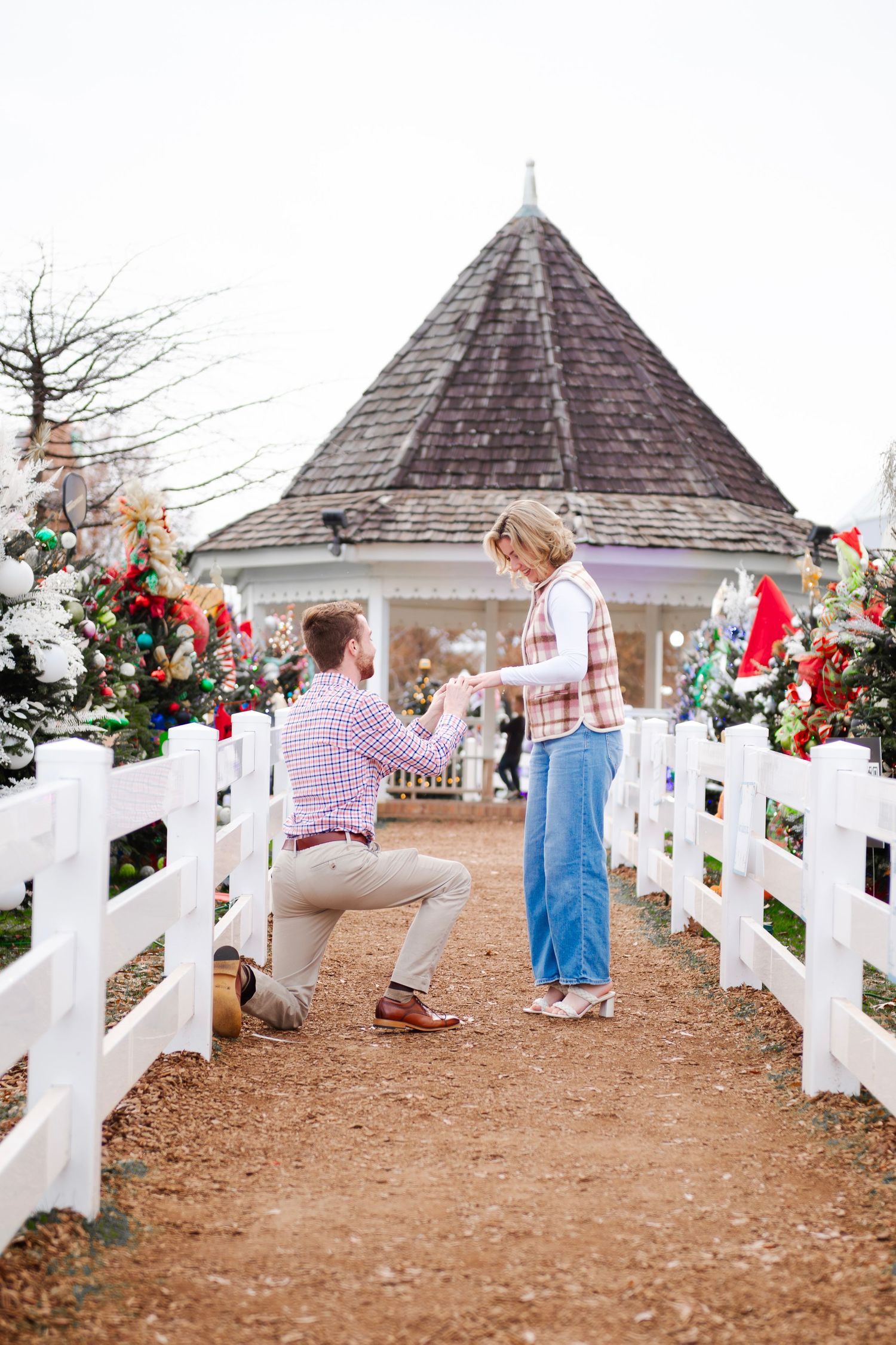 Romantic Christmas proposal with man on one knee before woman near a decorated gazebo with white fence.