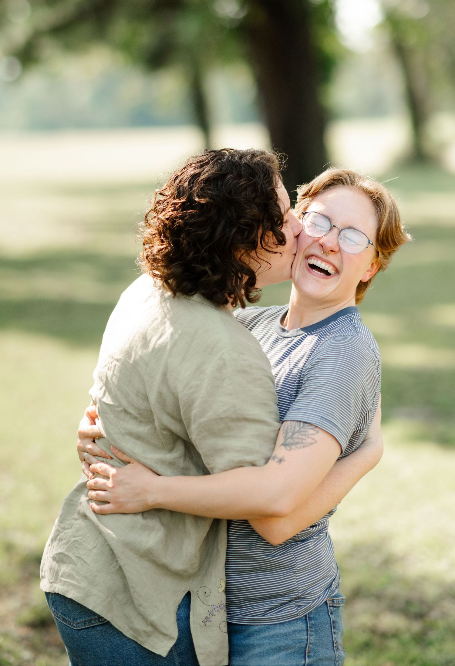 Two women laughing and hugging each other joyfully in a bright outdoor park setting.
