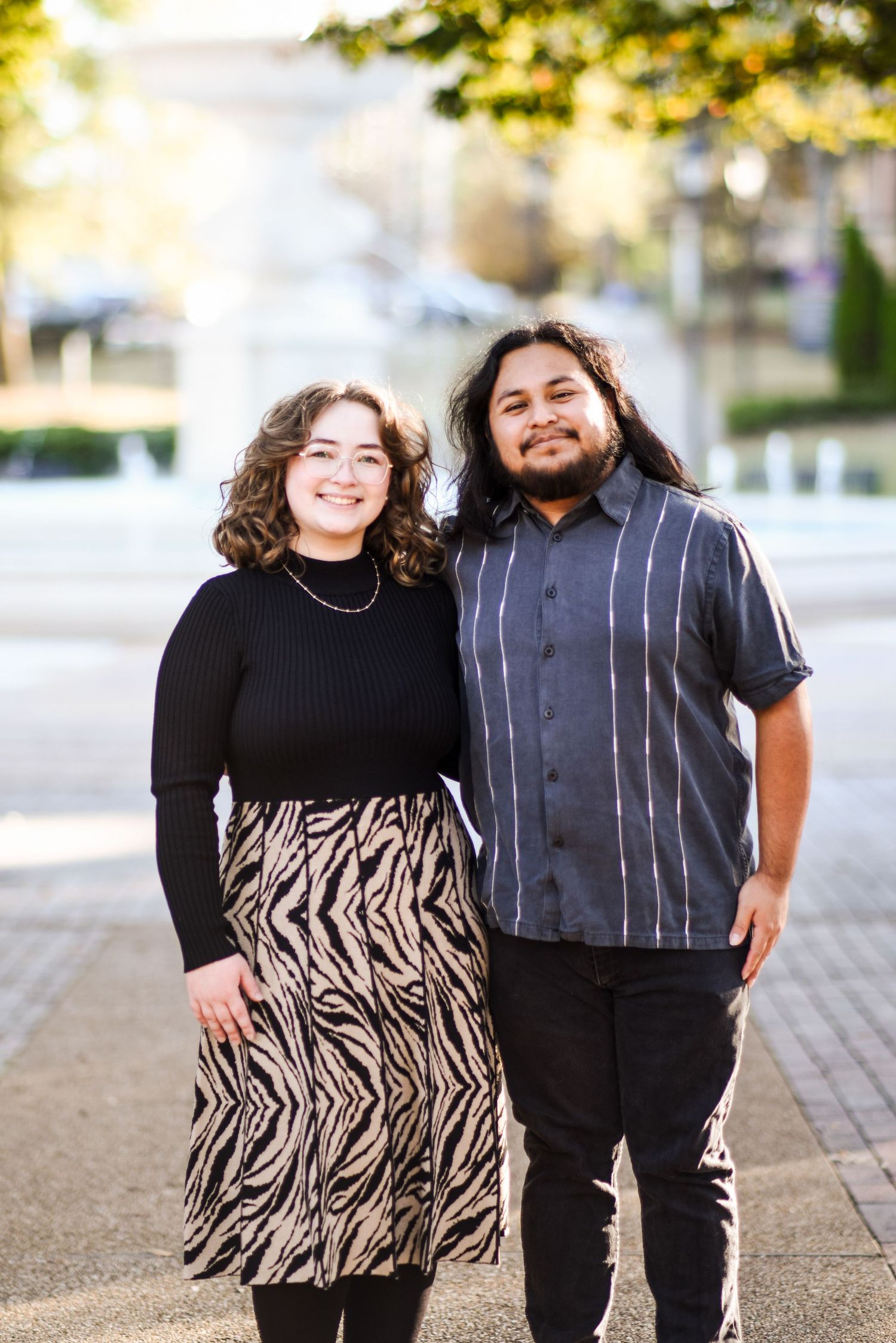 Smiling couple posing together on a sunny urban sidewalk, woman in black sweater and zebra skirt, man in striped button-up shirt.