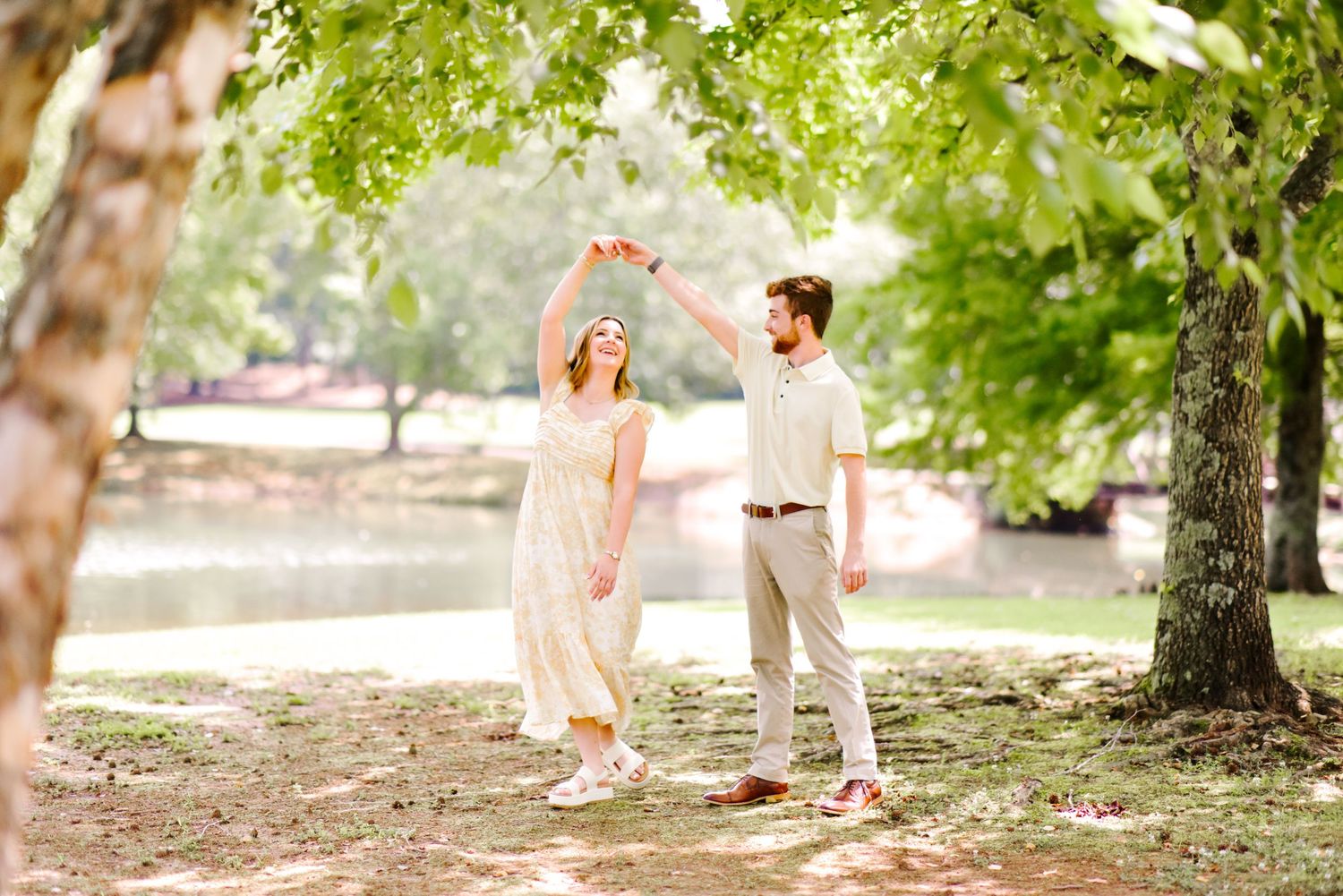 Couple dancing together under green trees near a lake in a sunny park, woman in white dress being twirled.