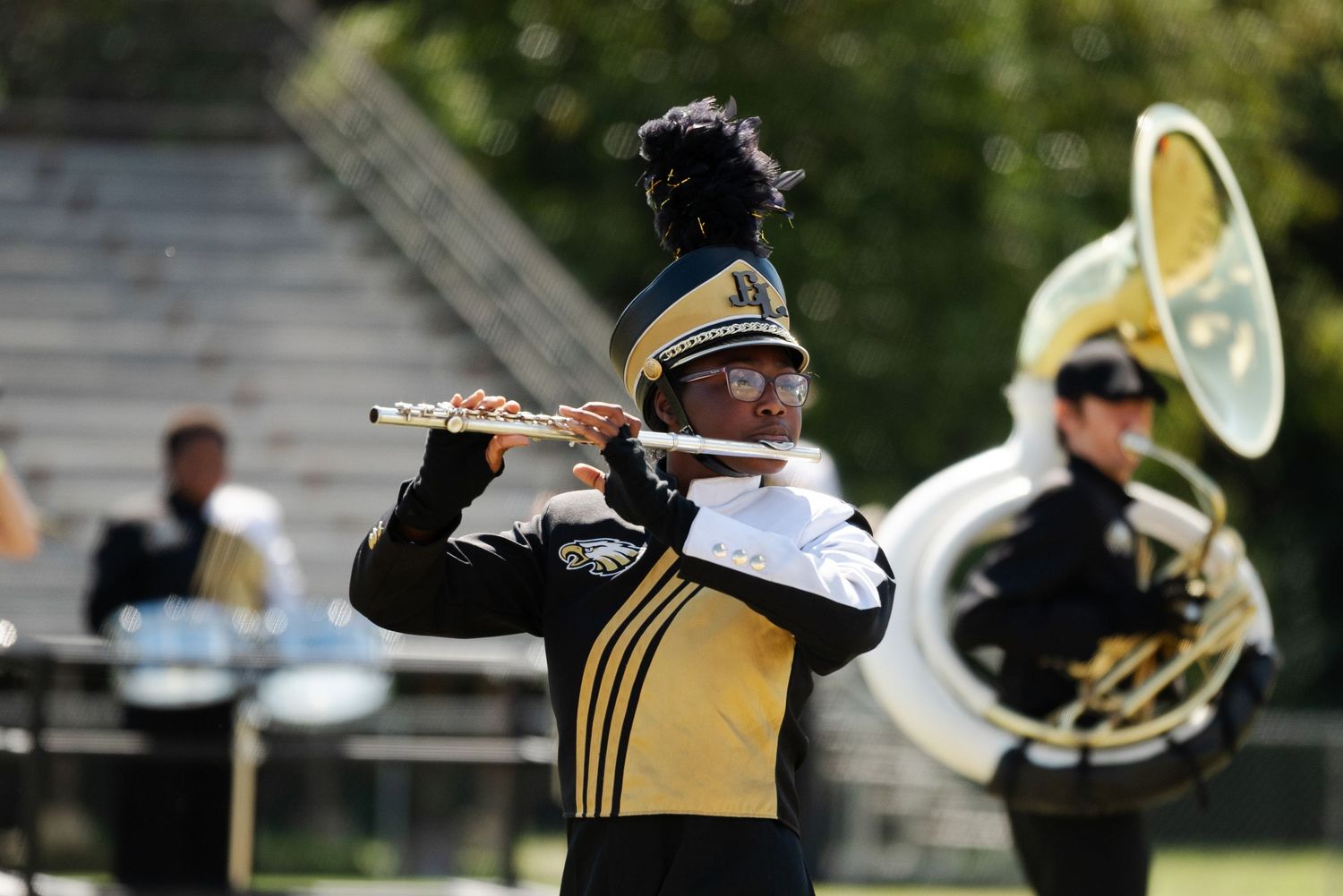 Marching band flutist in black and gold uniform performing on field.