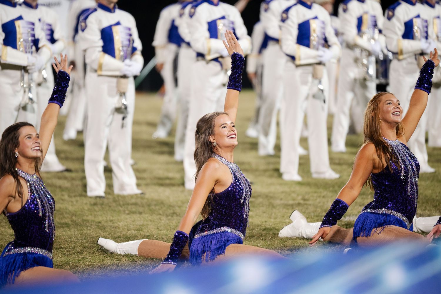 Smiling female dancers in blue sequined costumes perform on field during halftime show.