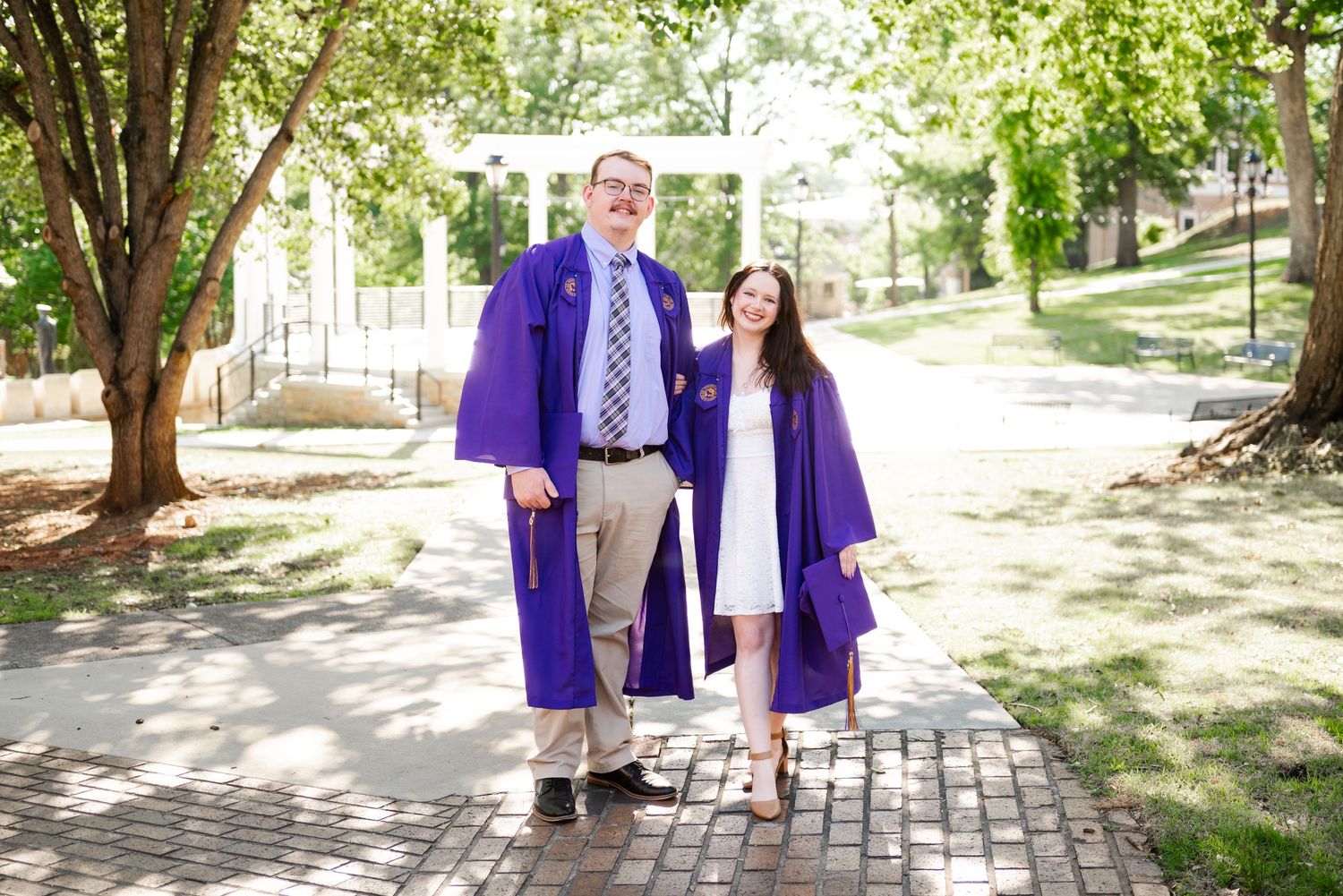 Two graduates in purple robes pose together on sunny tree-lined brick campus walkway.