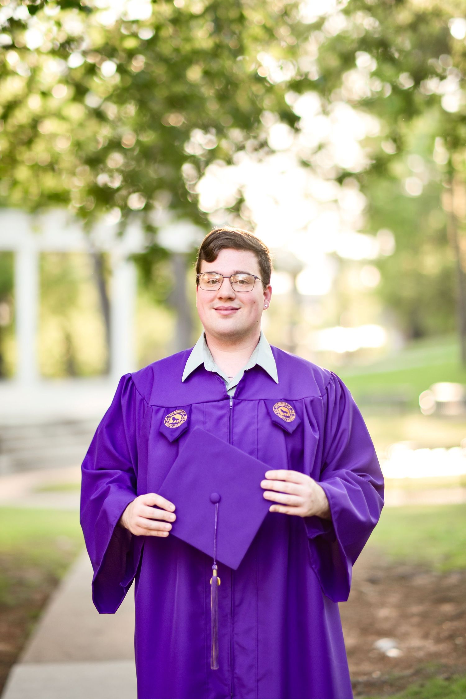 Young man in purple graduation gown holding cap outdoors among green trees.