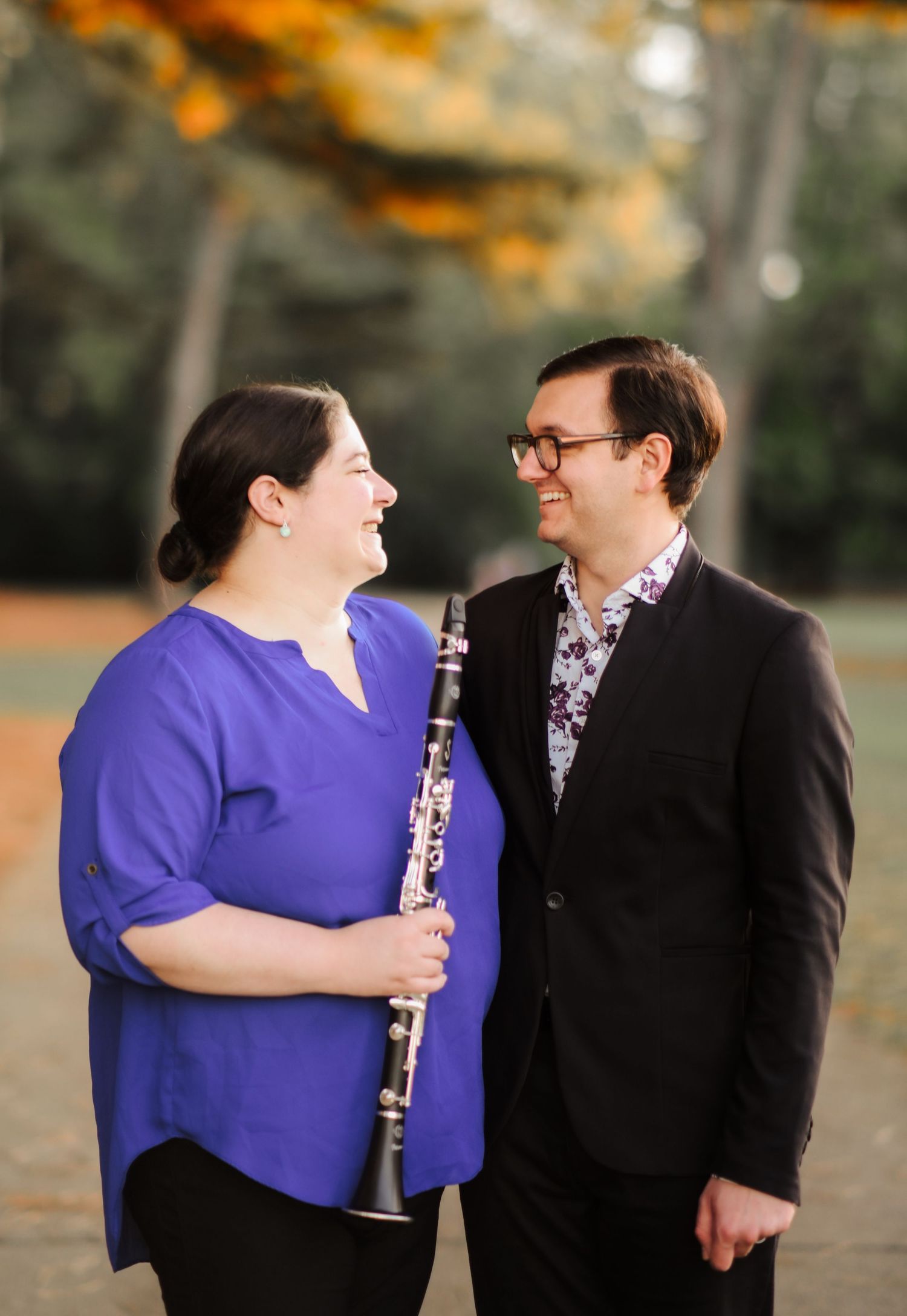 Couple smiling at each other in an autumn park, woman in purple holding a clarinet, man in black suit with floral shirt.