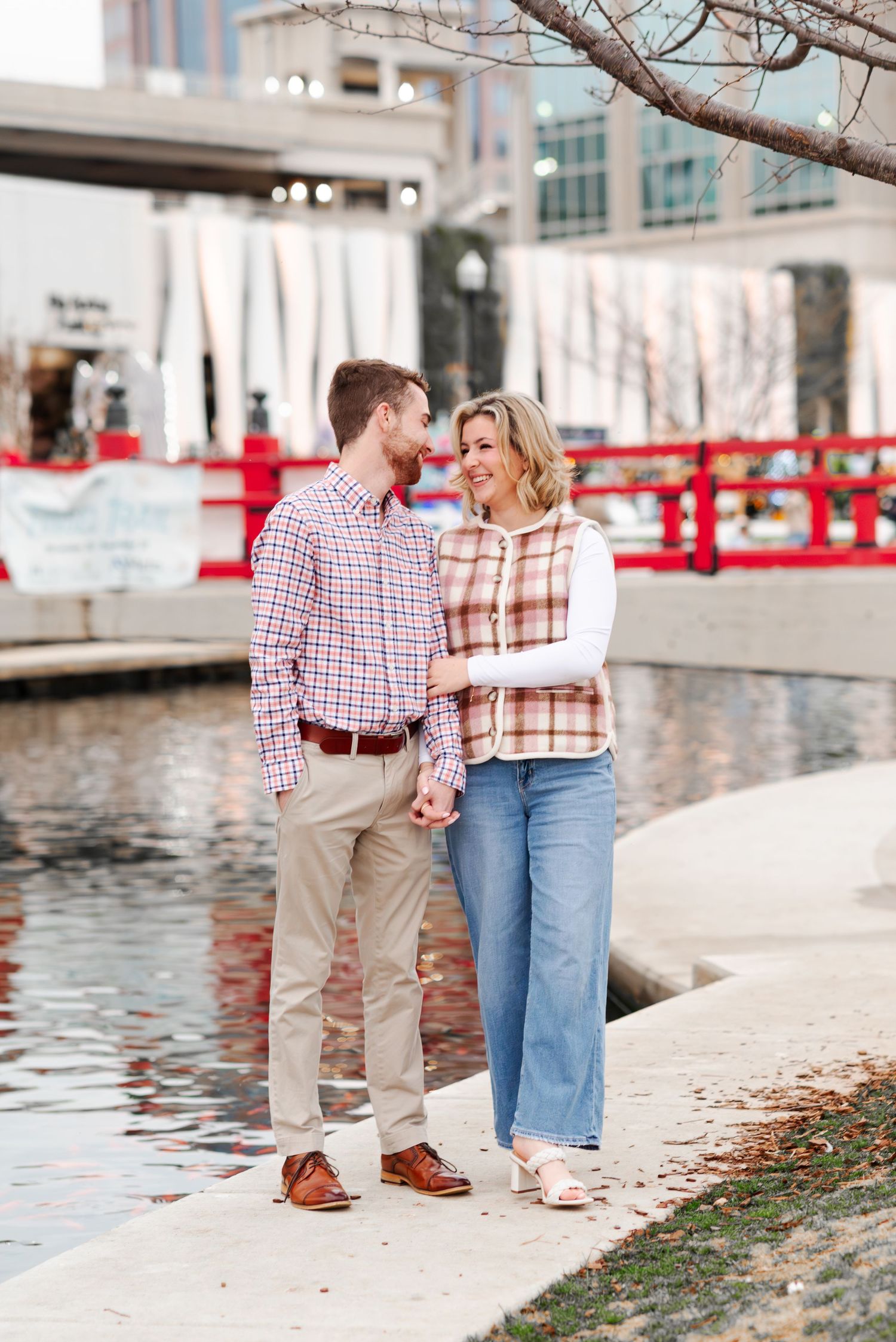 Couple holding hands and gazing at each other beside a reflective pond with a red bridge backdrop.