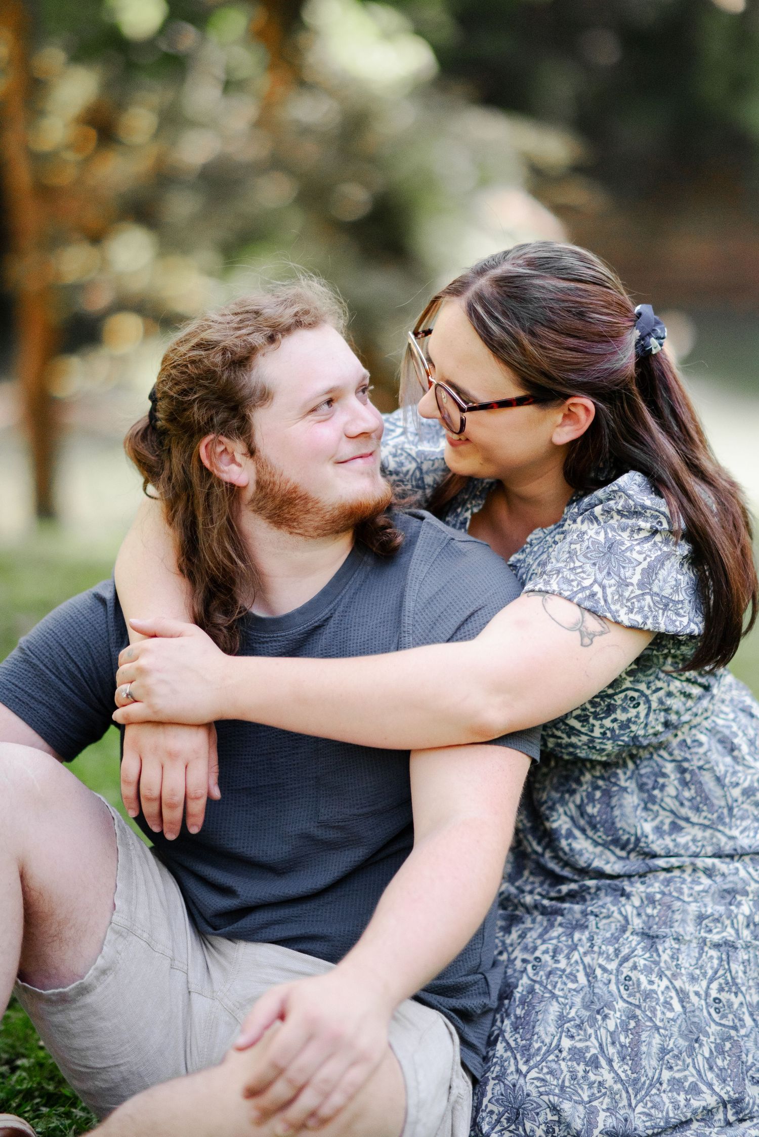 Couple sitting on grass embracing and smiling at each other in a bokeh outdoor setting.