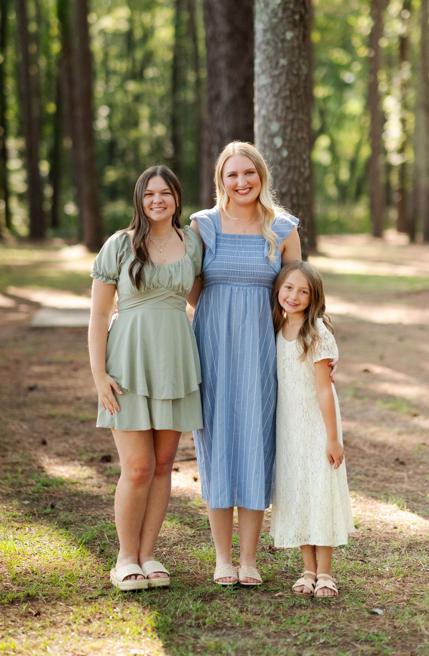Three sisters in green, blue, and white dresses smile together in a sunlit pine forest setting.