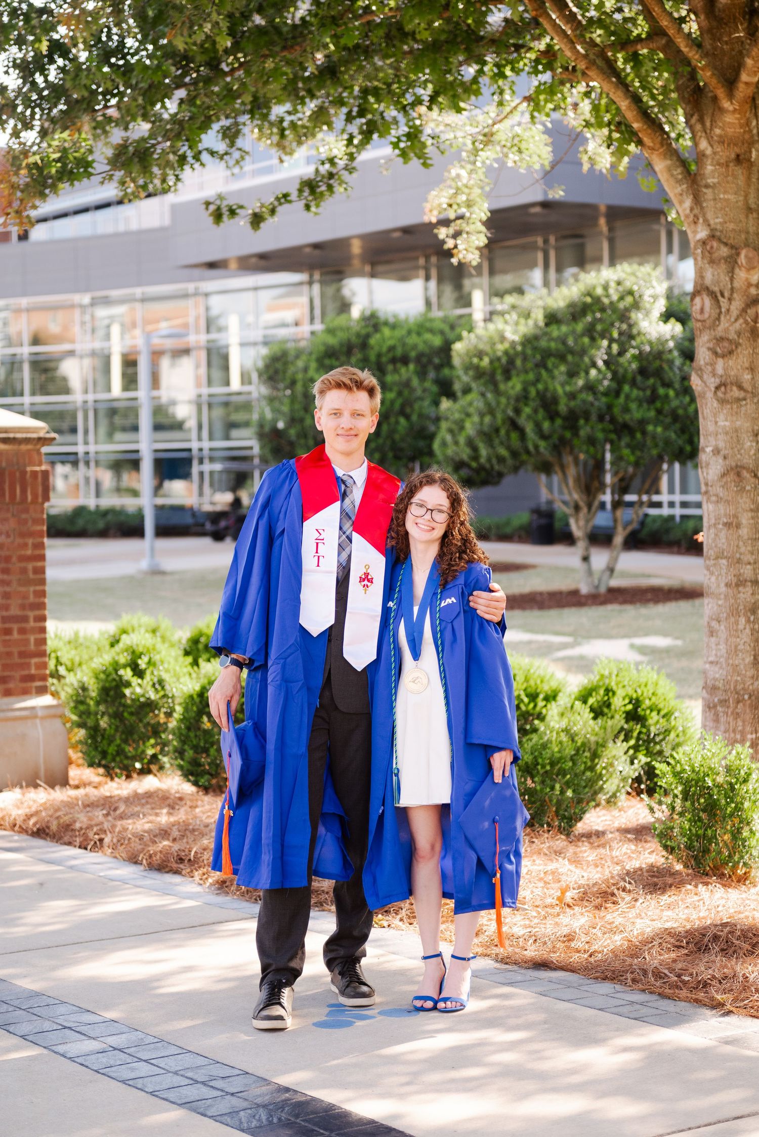 Two graduates in blue robes smiling together on a sunny campus pathway surrounded by trees.