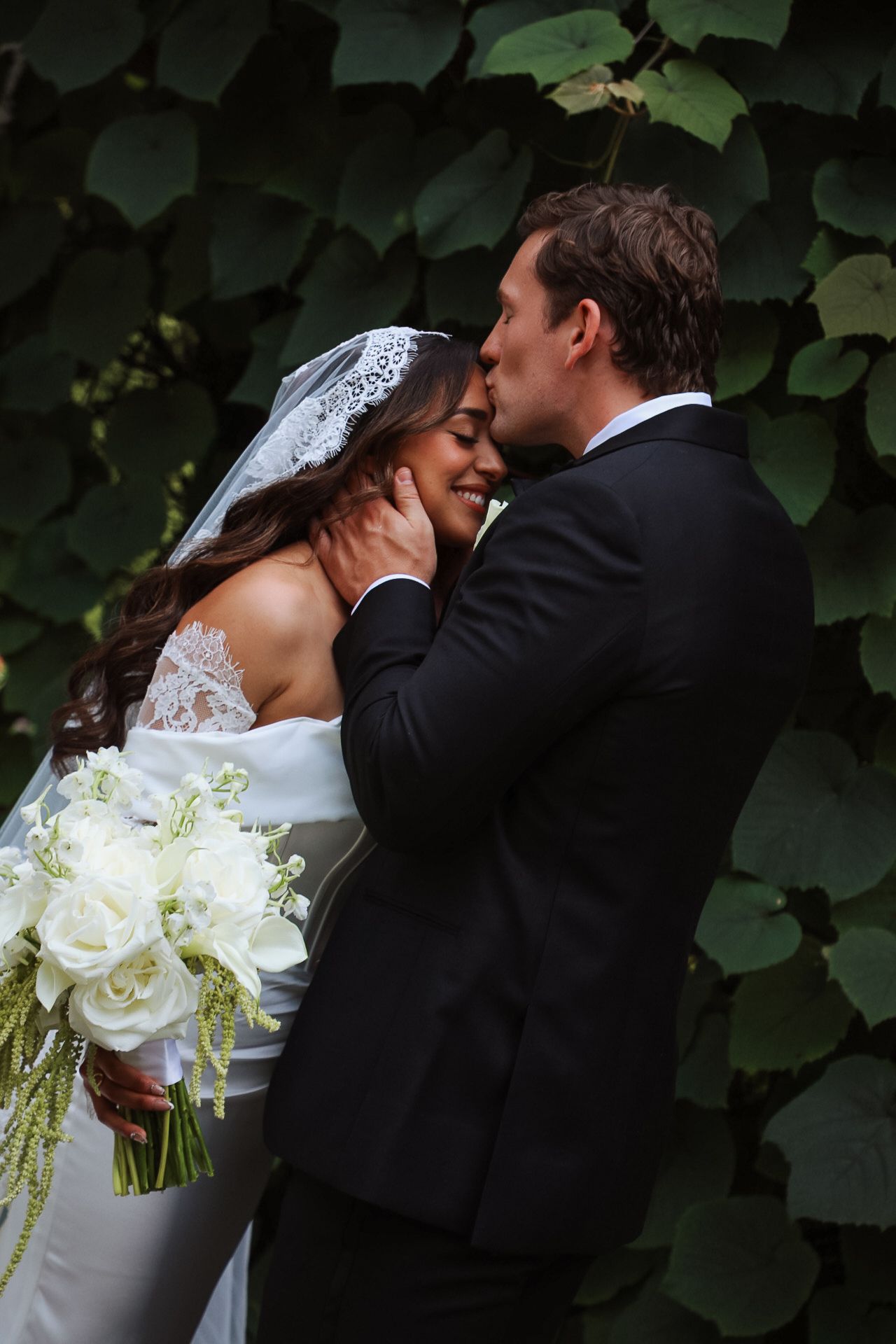 Bride and groom share an intimate embrace in front of lush green ivy wall, bride holding white rose bouquet.