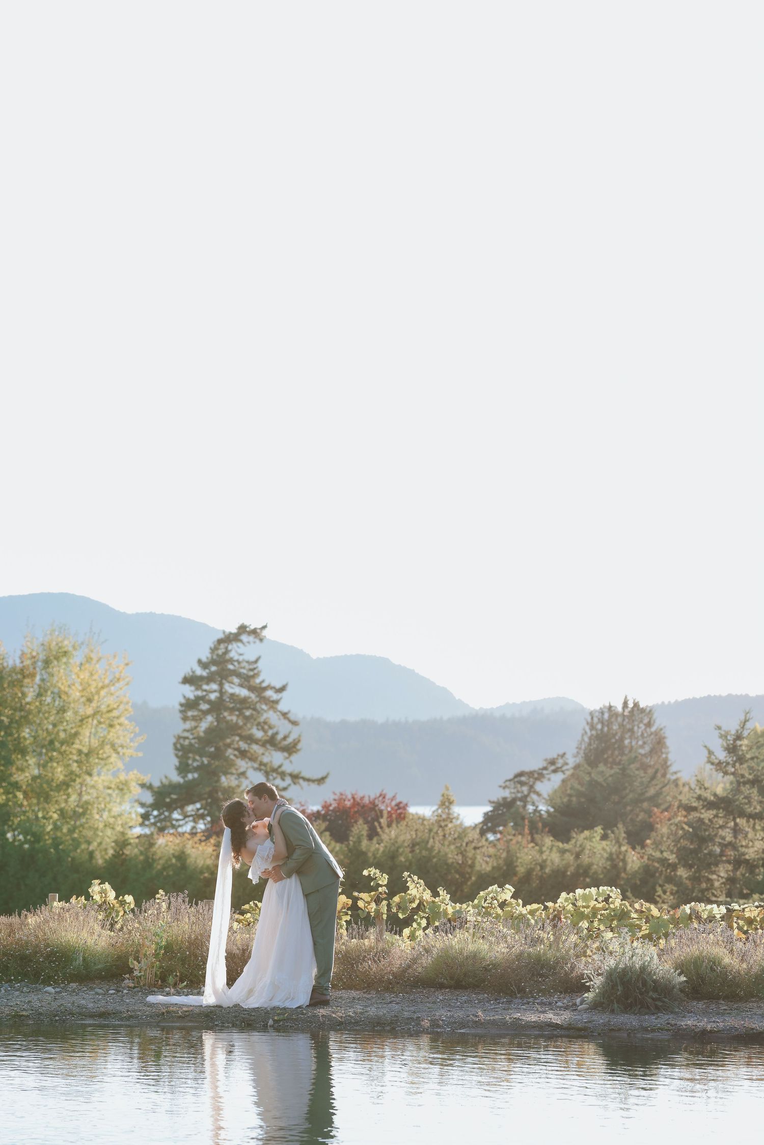 Bride and groom share a romantic kiss by a serene lake with mountains and trees in the background.
