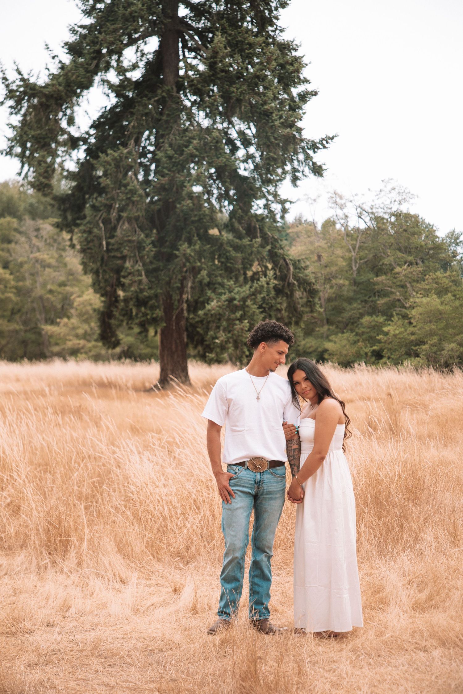 A couple stands in a golden field, the man in white tee and jeans, the woman in a flowing white dress, with evergreen trees behind.