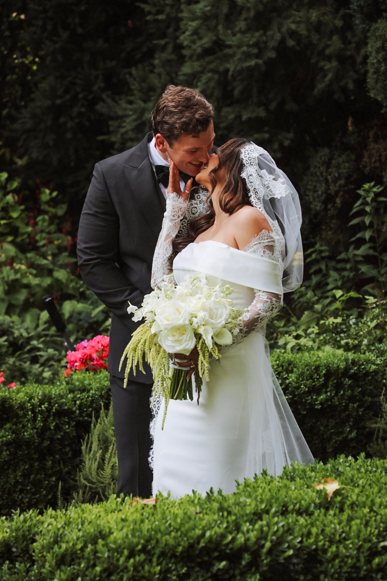 Bride and groom kissing in lush garden, bride holding white bouquet with lace veil and off-shoulder gown.