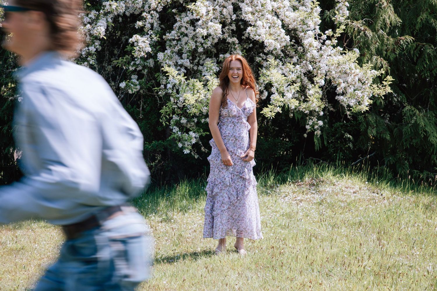 Woman in floral dress smiles in a sunlit garden with blooming white trees in the background.