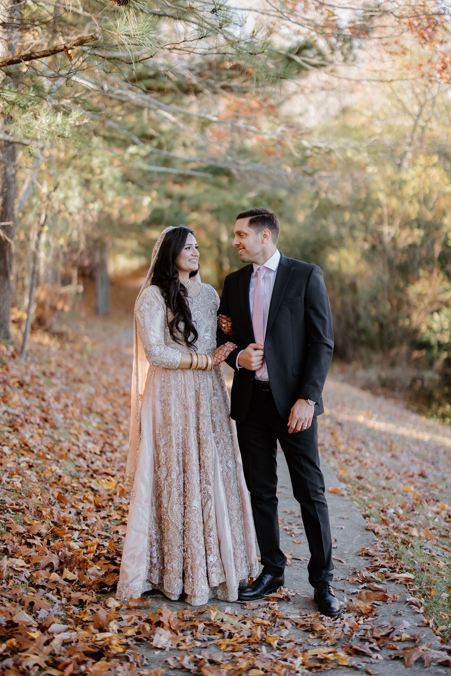 A couple in formal attire poses together on a path covered in autumn leaves.