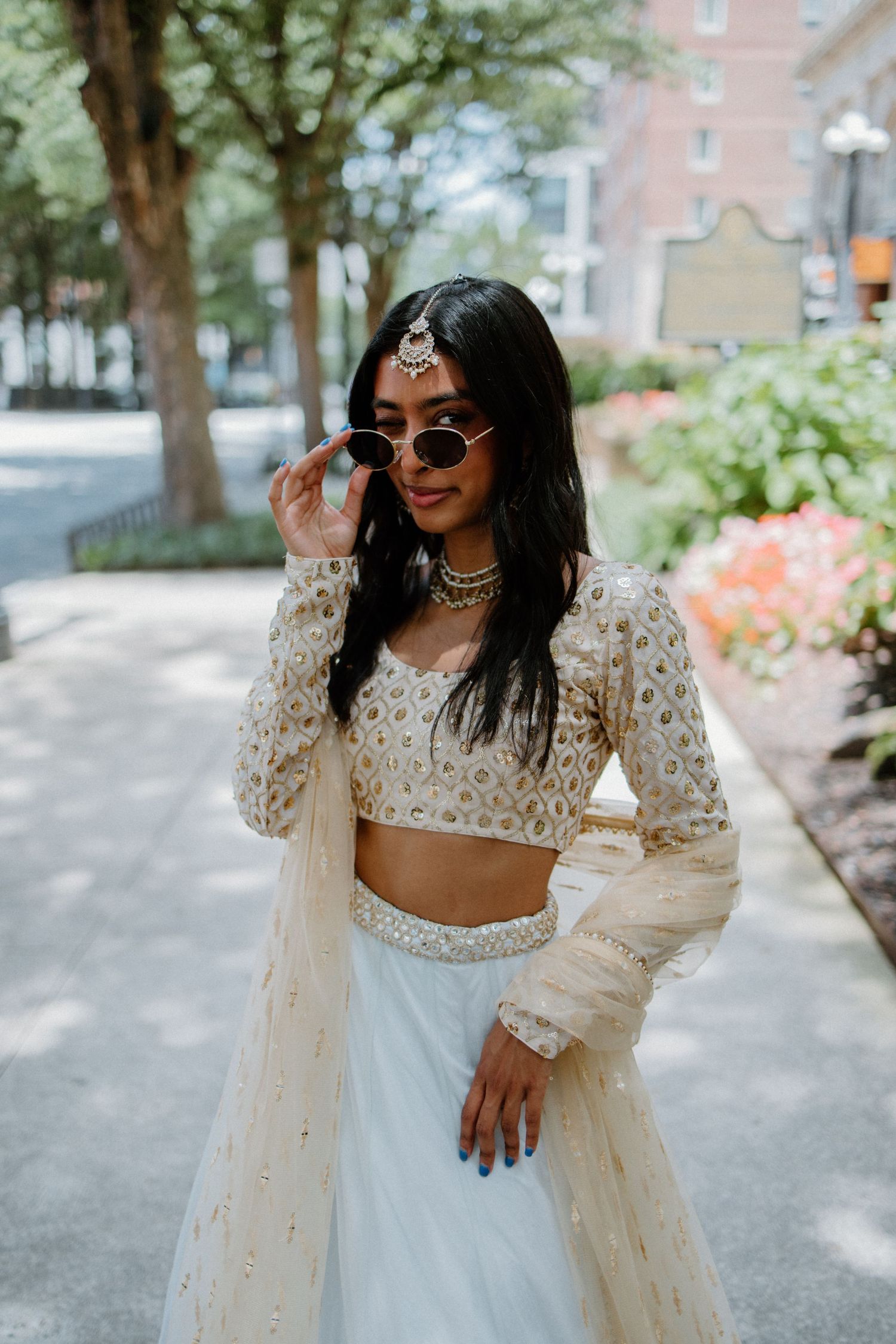 Stylish woman in white crop top and full skirt adjusts sunglasses while posing on tree-lined city street.