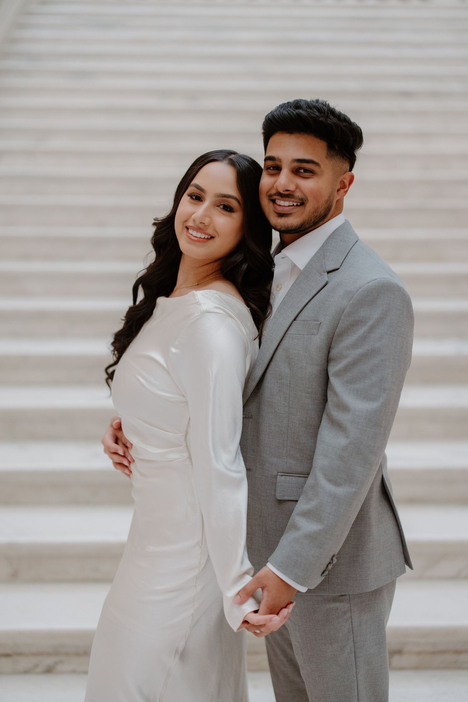 A couple in wedding attire pose on white steps, with the bride in a sleek white dress and groom in a light grey suit.