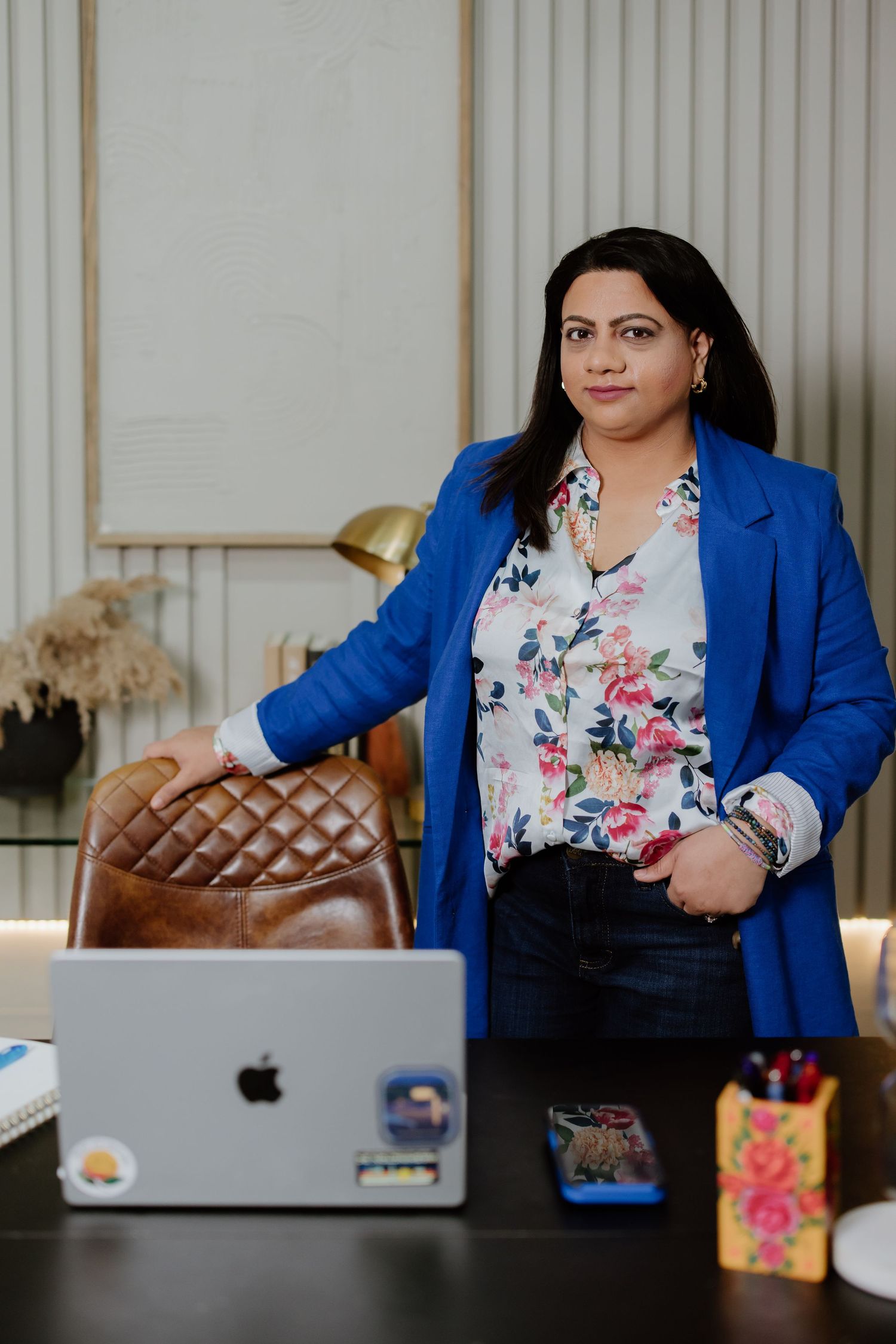 Professional in a bright blue blazer and floral blouse stands at desk with laptop and office accessories.