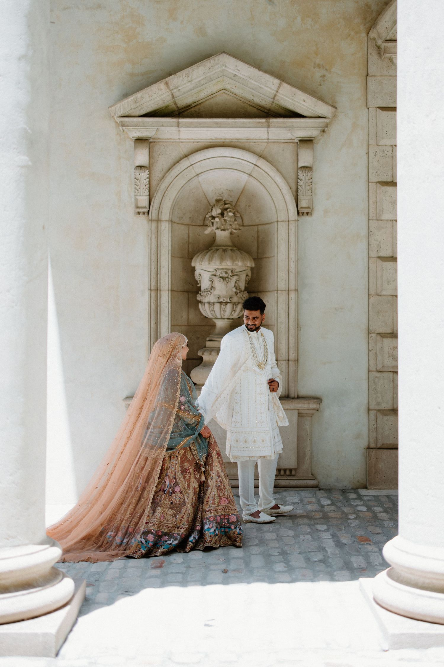 A bridal photoshoot with flowy peach tulle gown against ornate white stone columns and arched architecture.