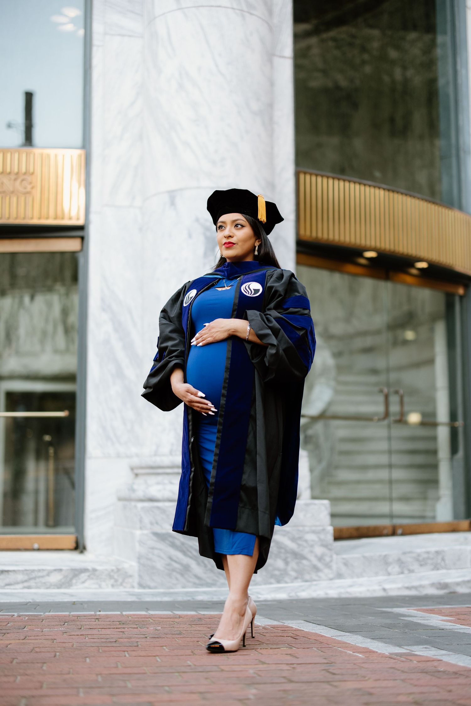 An expectant graduate stands in academic regalia including a blue and black graduation gown and cap outside a building.