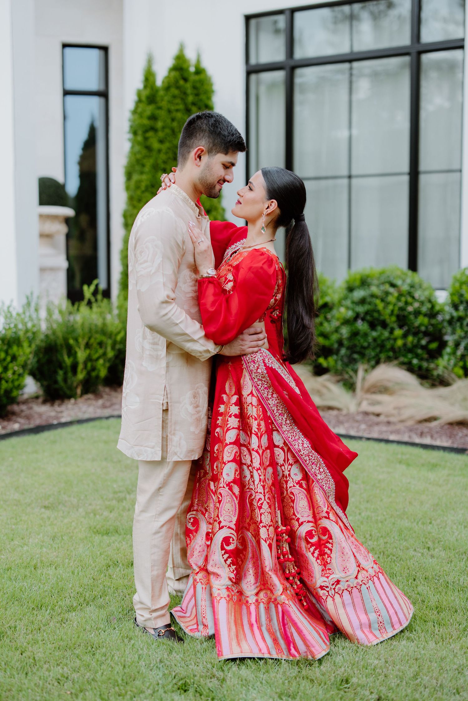 A romantic couple in red and beige traditional wedding outfits share a tender moment on a grassy lawn.