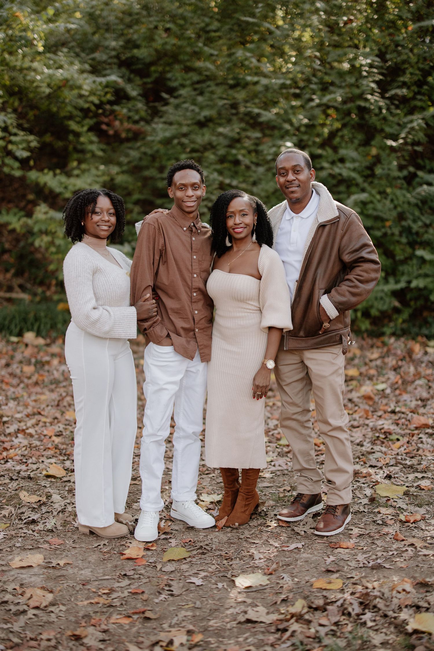 Family wearing coordinated brown and white outfits poses together on a fall day surrounded by autumn foliage.