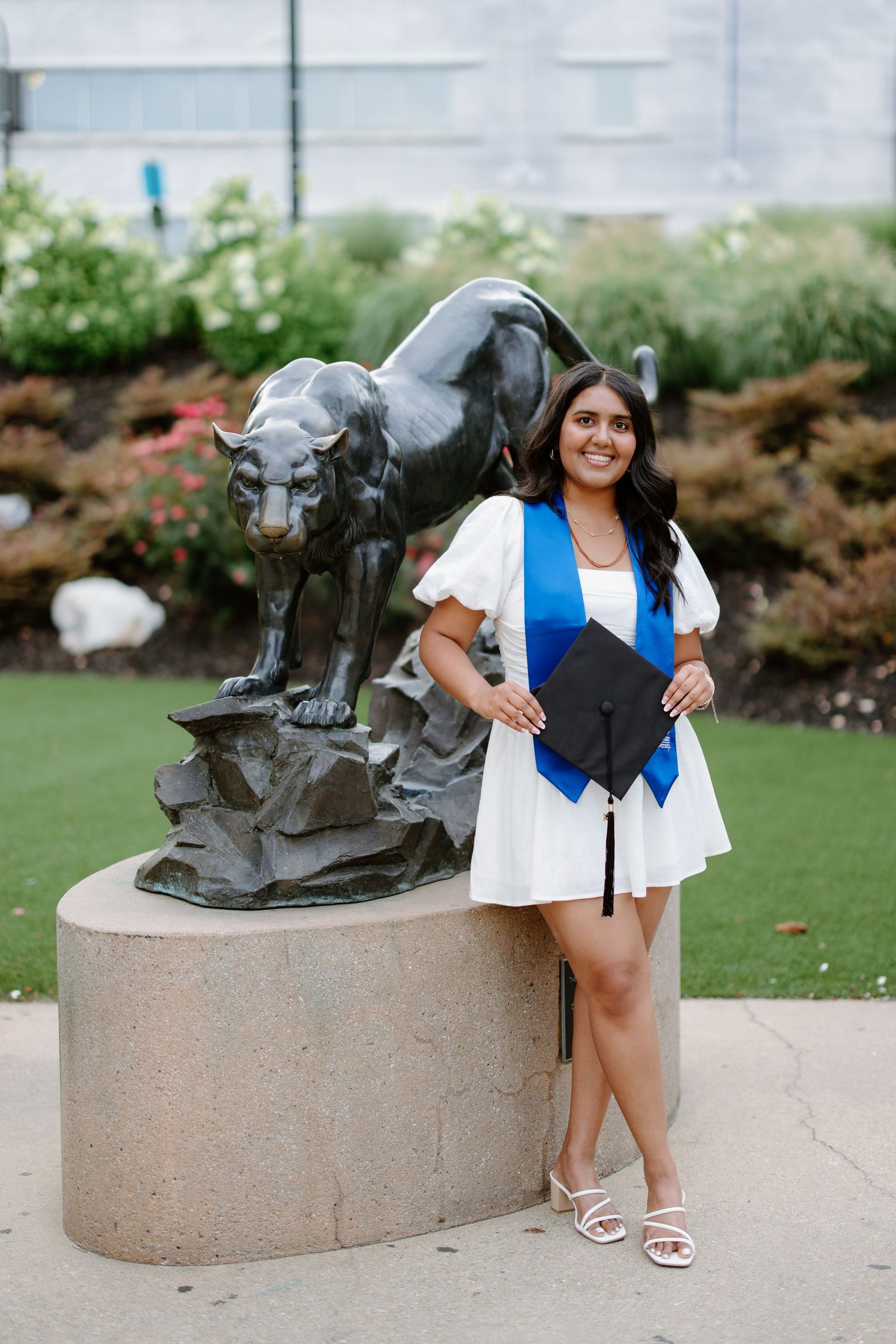 A graduate poses next to a large bronze panther statue on a concrete pedestal in an outdoor garden setting.