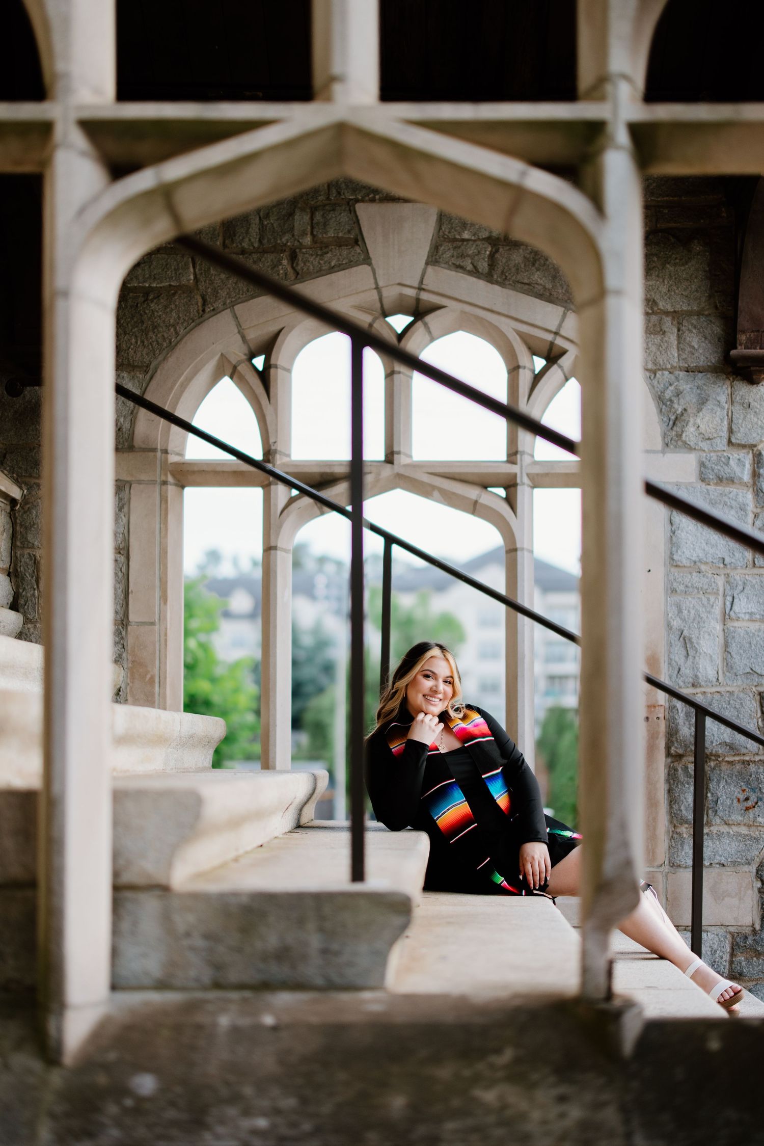 Graduate sits in a Gothic stone archway wearing academic regalia and honor cords during graduation photo session.