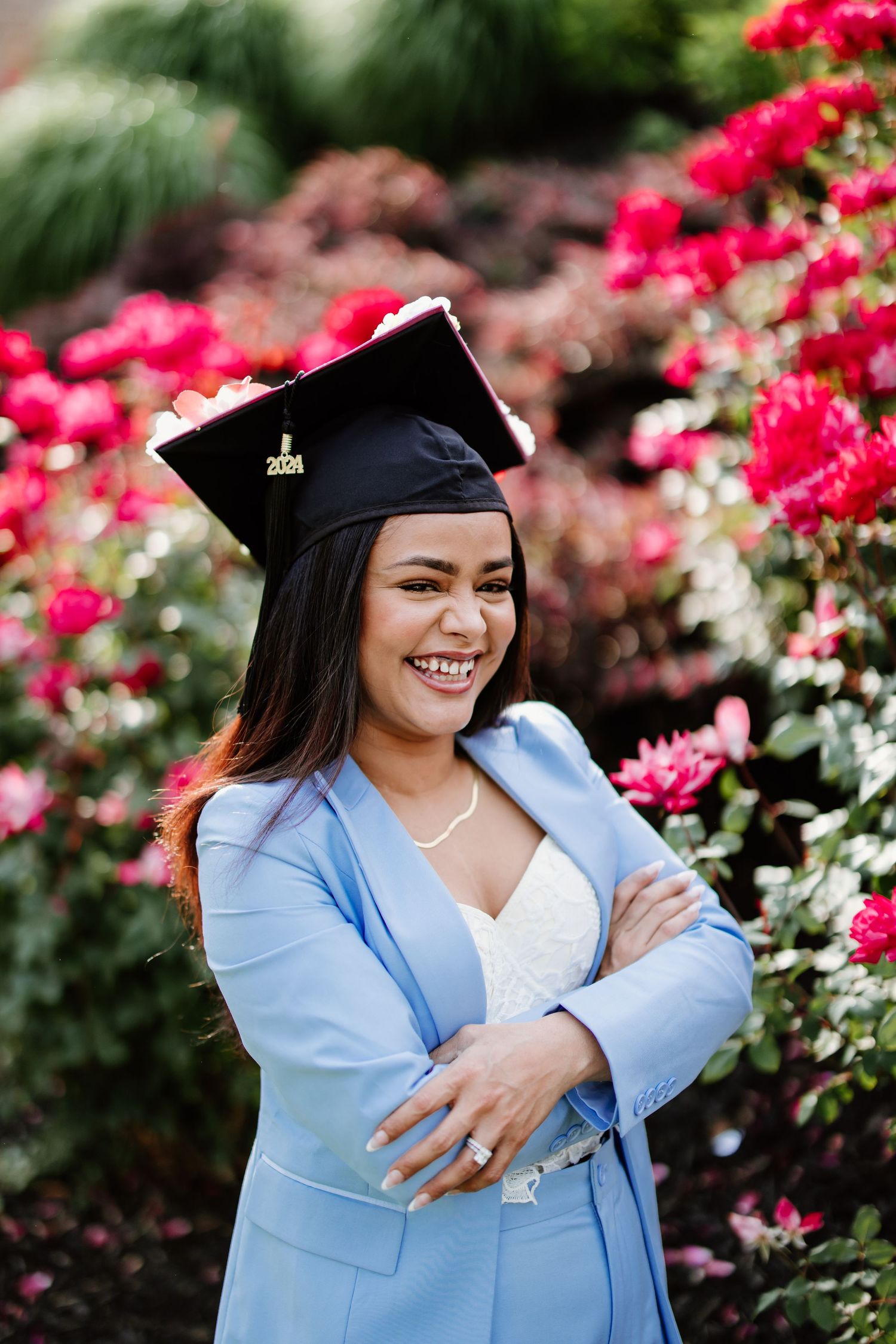 A graduate in light blue blazer and cap stands smiling in front of vibrant pink azalea flowers.