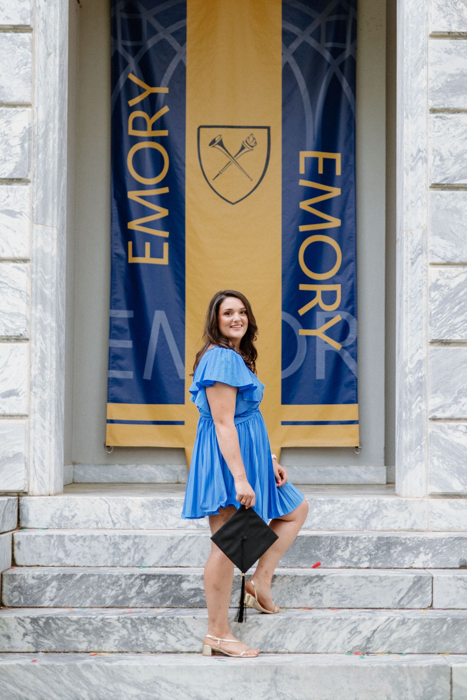 Graduate in blue dress poses on marble steps in front of Emory University banner displaying crossed keys shield emblem.