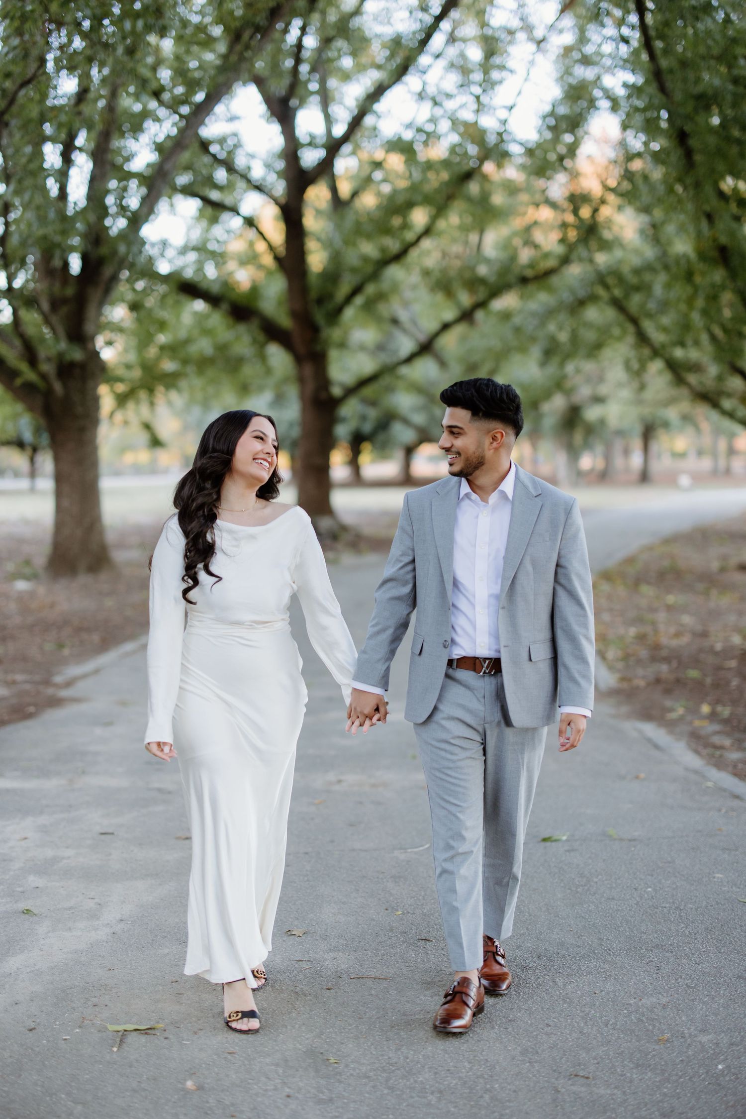 Couple in white dress and gray suit lovingly walk hand in hand down a tree-lined path.
