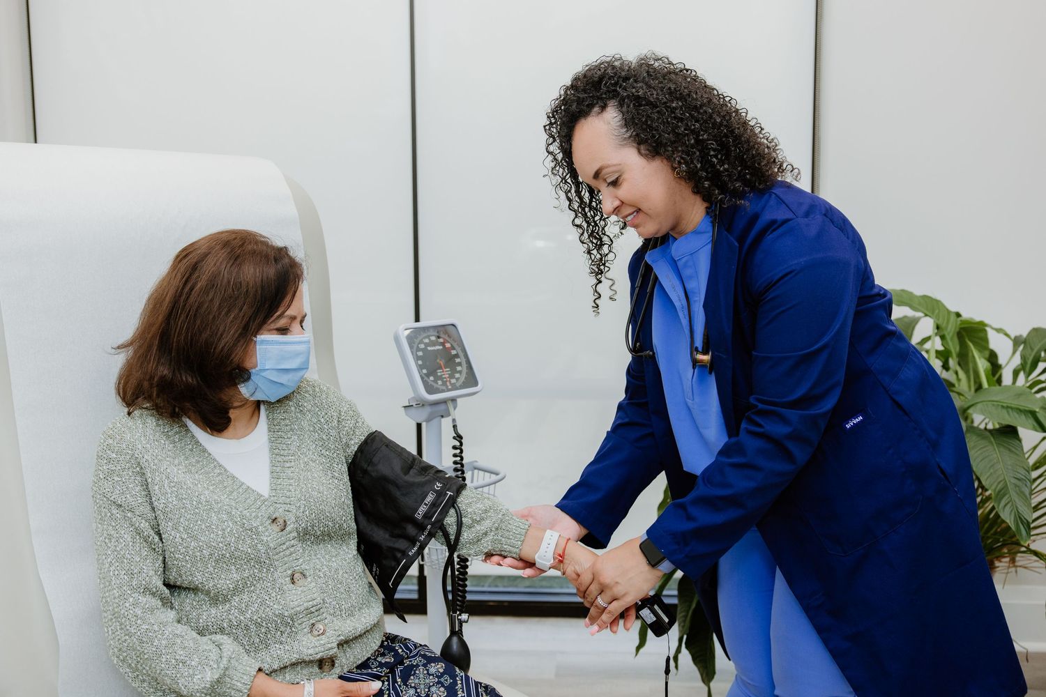 Healthcare professional in blue blazer checks patient's vitals while patient sits in medical exam chair wearing mask.