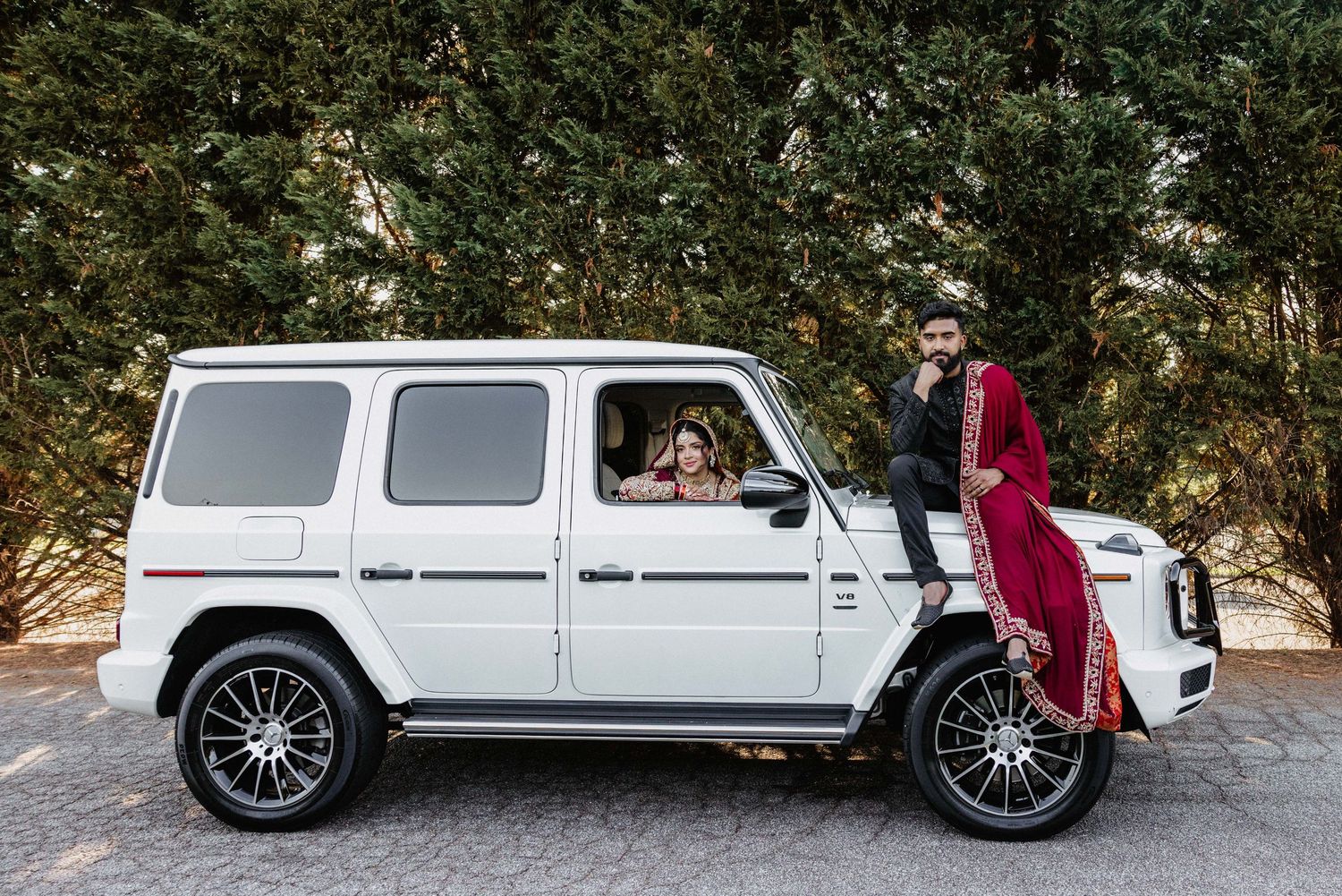 A white Mercedes G-Wagon luxury SUV parked on a road with someone wearing a red dress beside it.