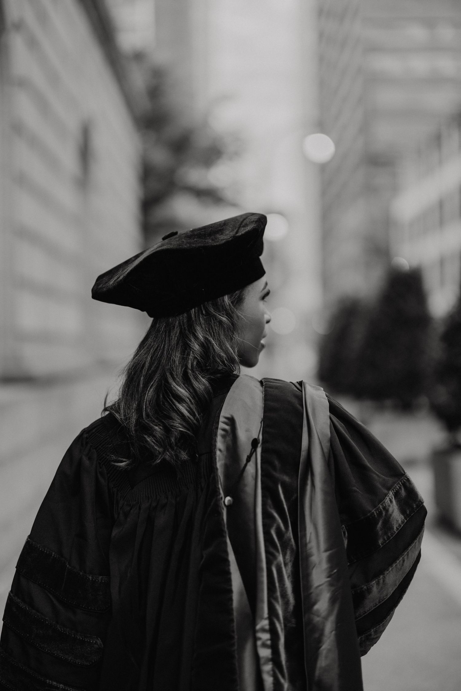 A graduate in black academic regalia looks away, photographed in black and white on a city street.