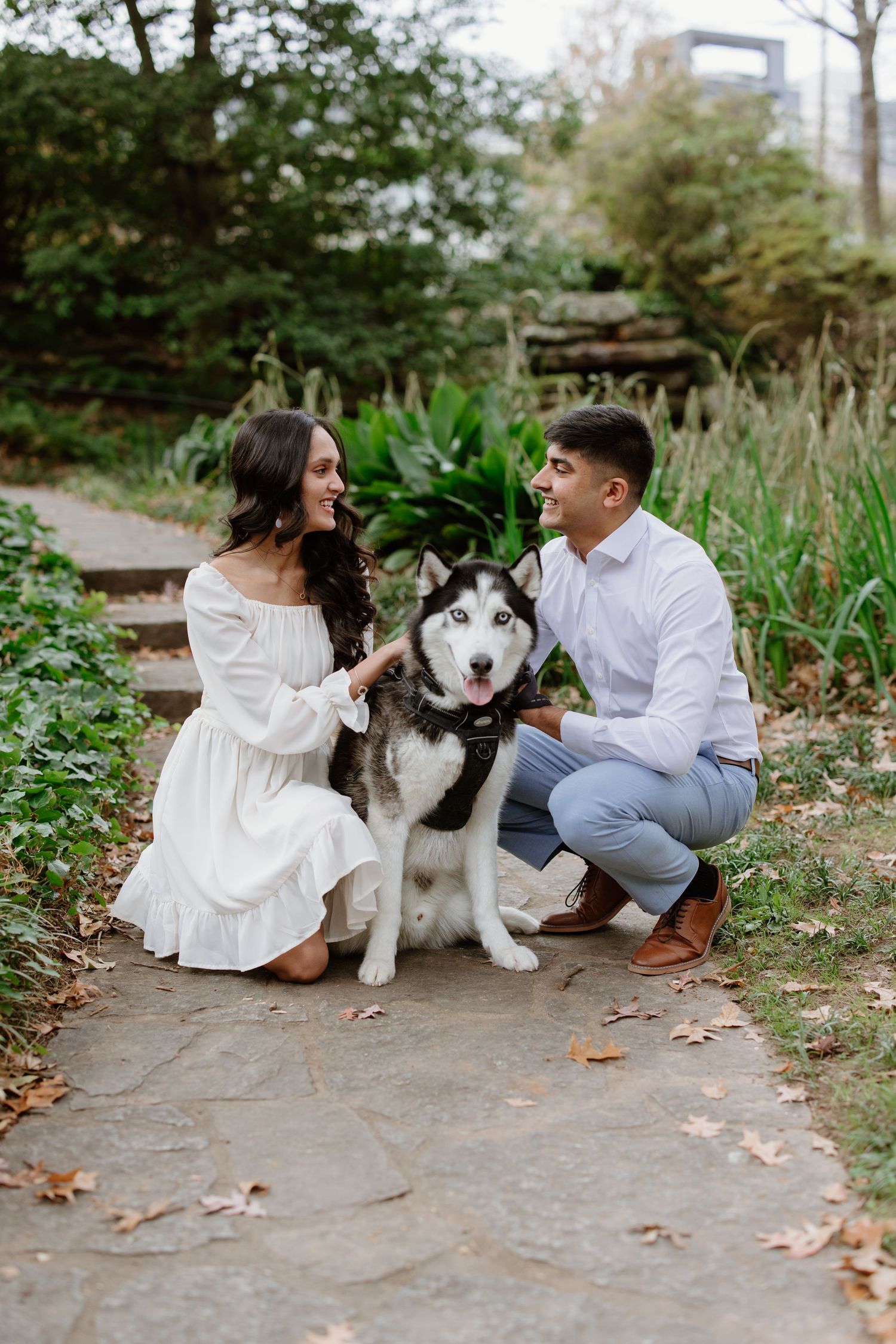 A couple sits on a path with their husky dog during an outdoor photo session in a natural setting.