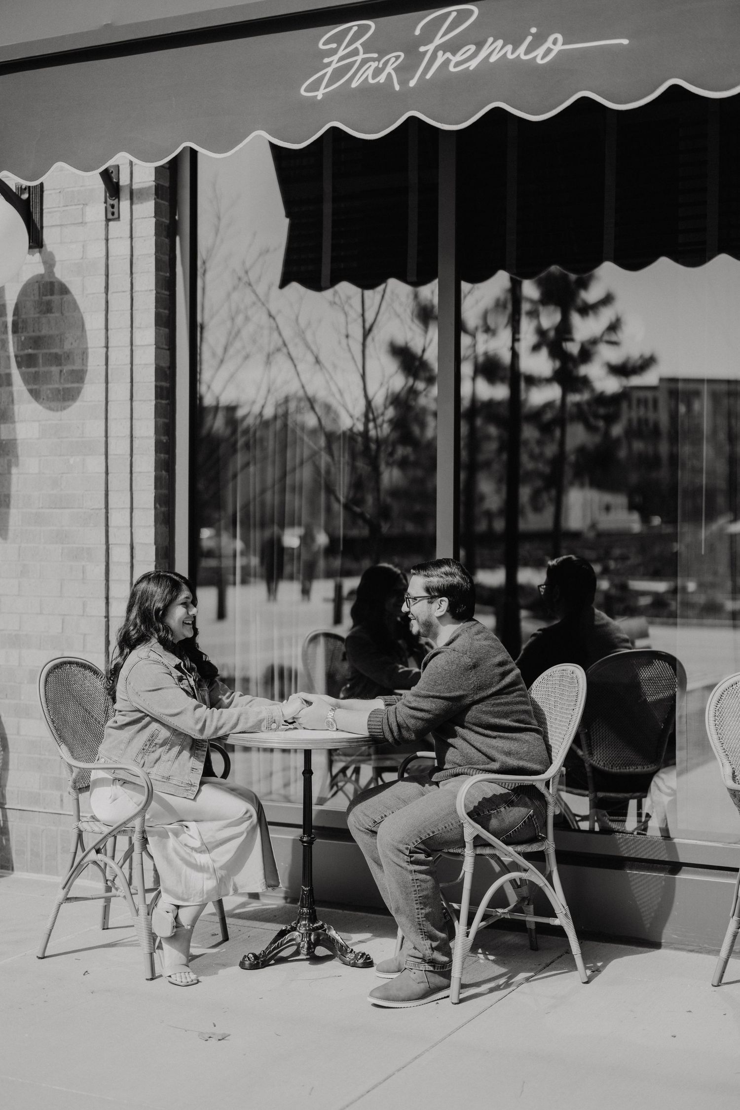A black and white photo showing people seated at an outdoor cafe table under an awning on a sunny day.