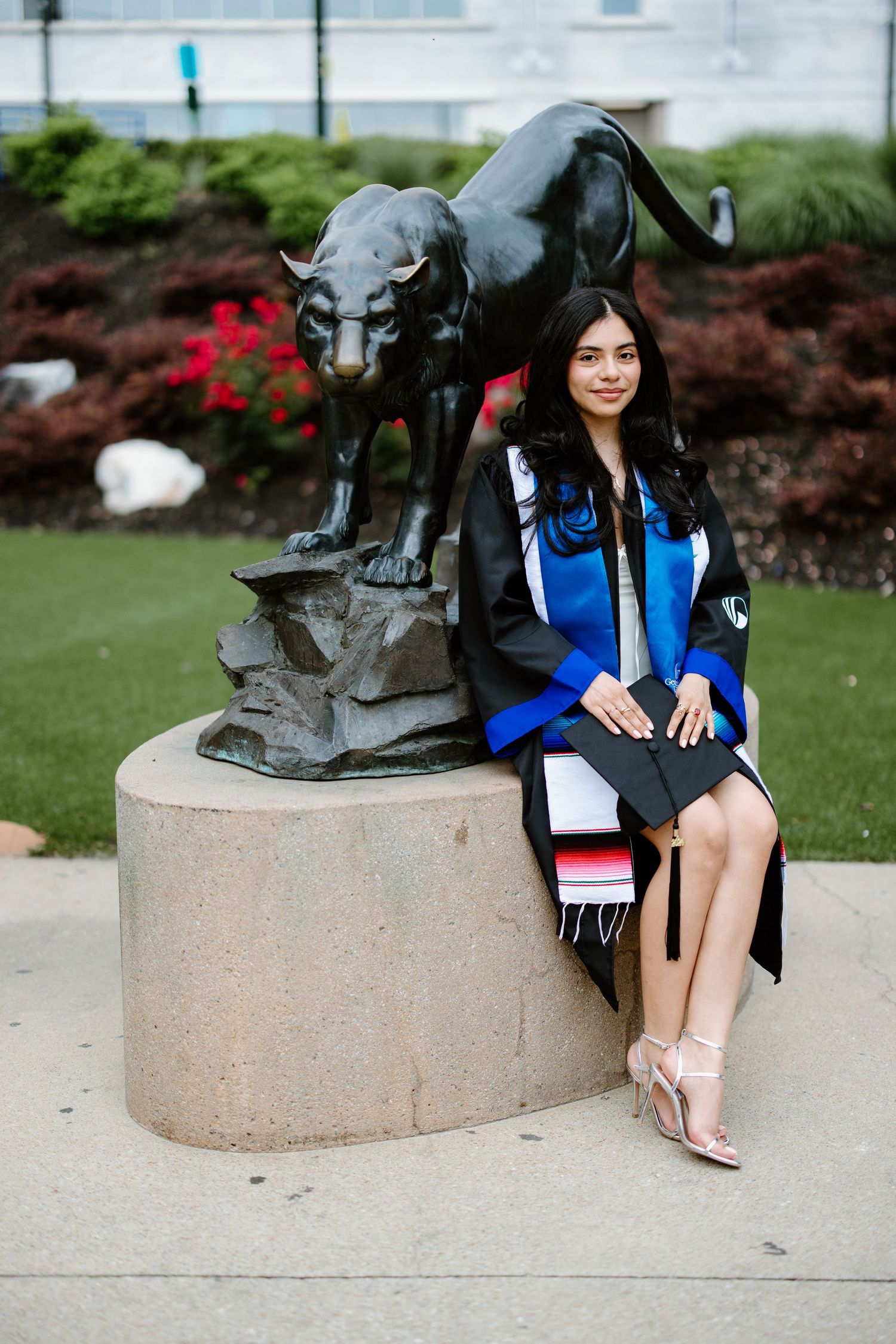A bronze panther statue stands on a concrete pedestal next to a graduate in academic regalia on a grassy campus lawn.