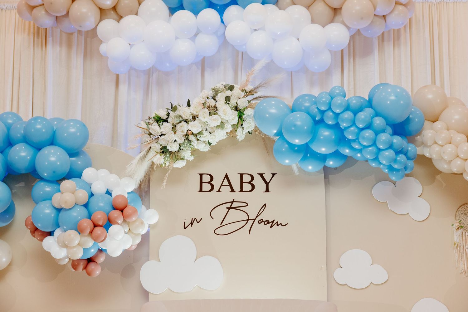 A baby shower decoration setup with white and blue balloon clouds and garlands against a beige wall.