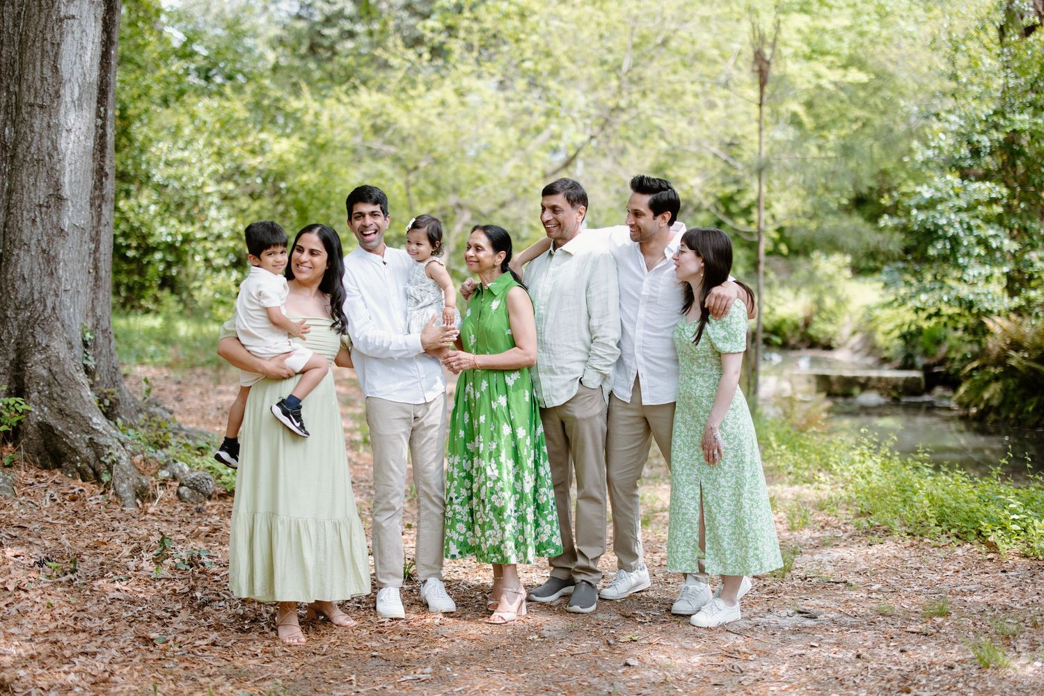 A wedding party wearing green and white attire poses together in a scenic outdoor woodland setting.