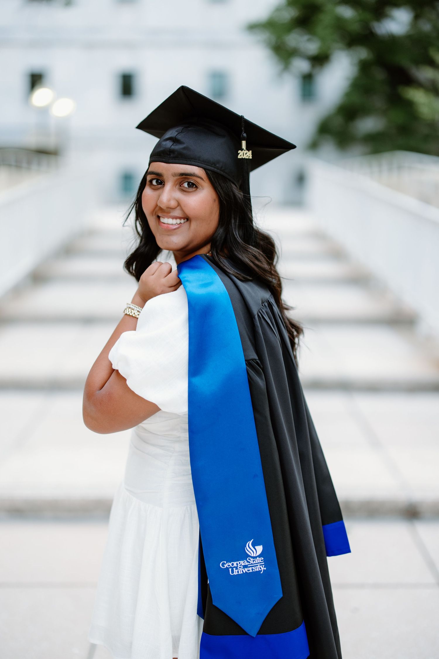 A graduate poses in white dress and black graduation cap and gown with blue sash during outdoor portrait session.