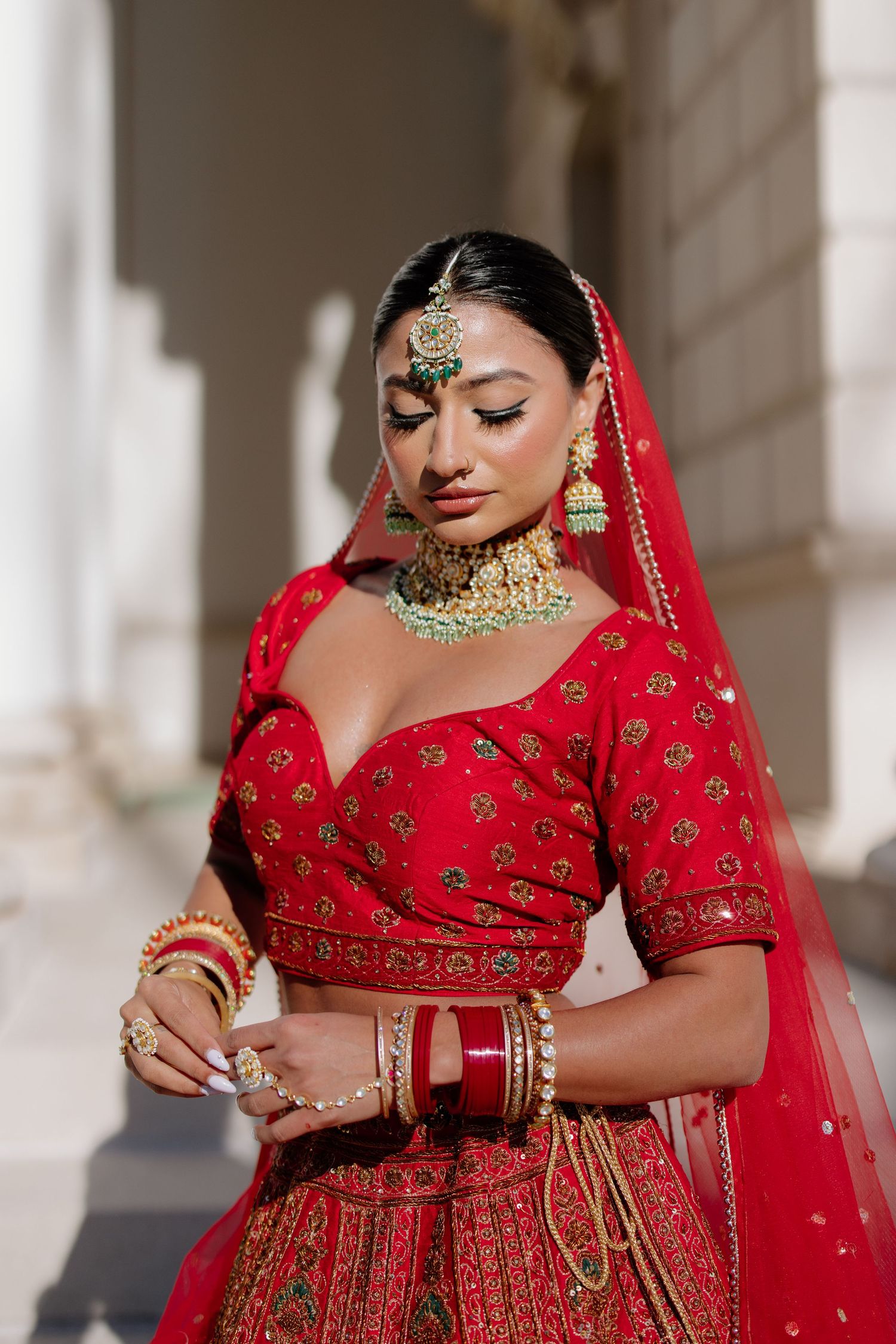 Beautiful Indian bride in traditional red lehenga and gold jewelry poses elegantly against white pillars.