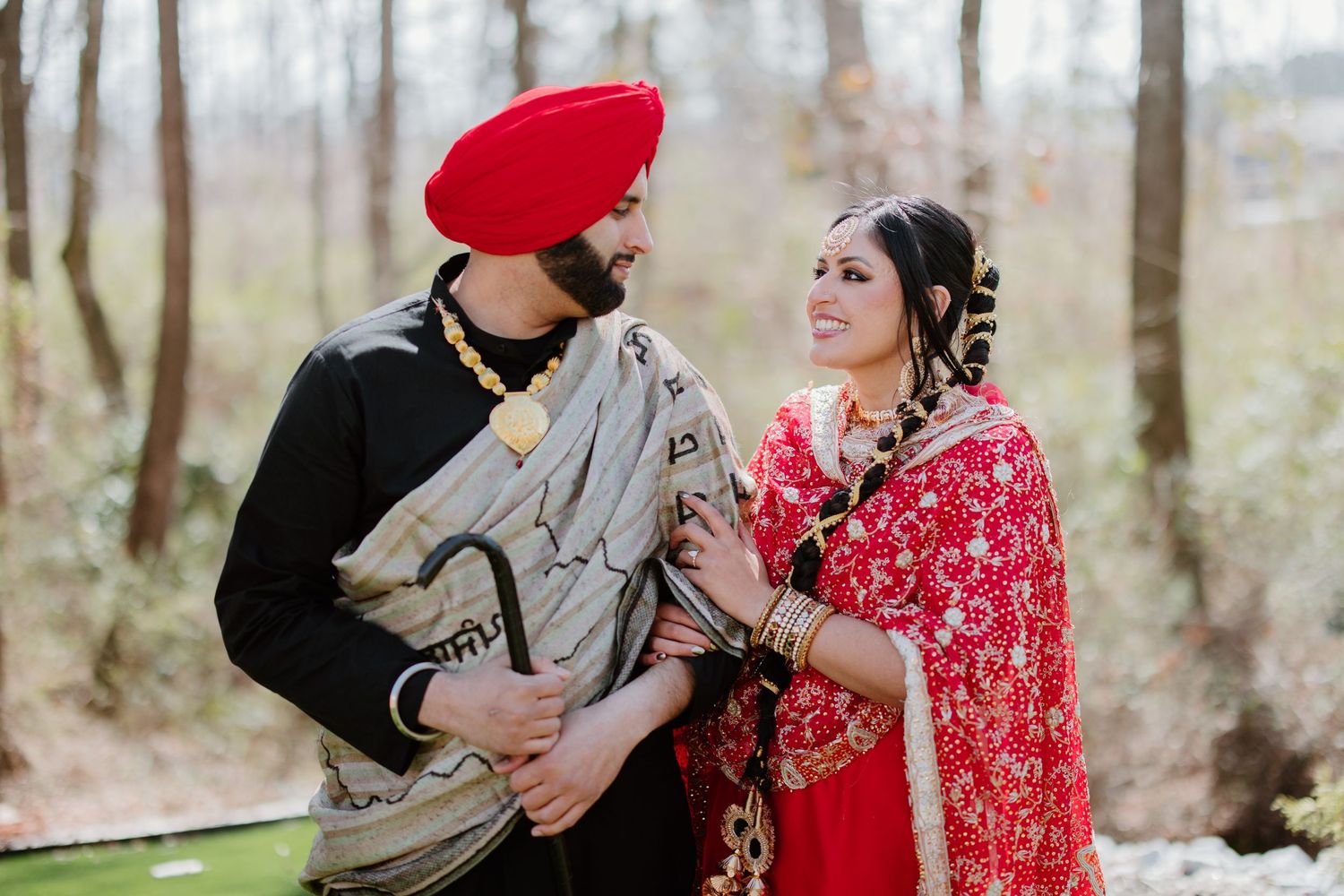 A couple in traditional Indian attire share a tender moment outdoors, with her in a red sari and him in a turban.