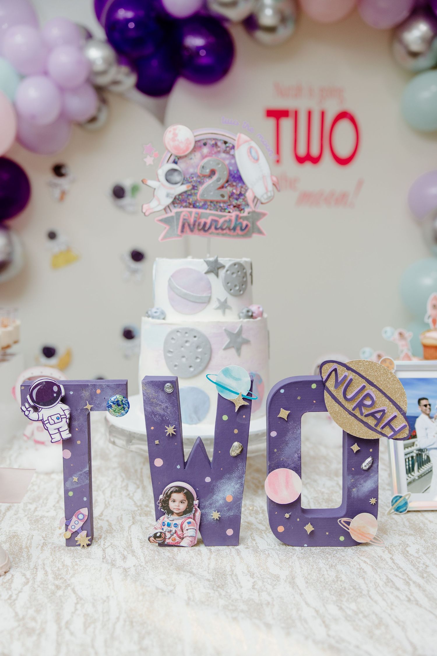 Purple and pastel balloon arch decorates a celestial-themed 'TWO' display with sparkly letters and space-themed decorations.