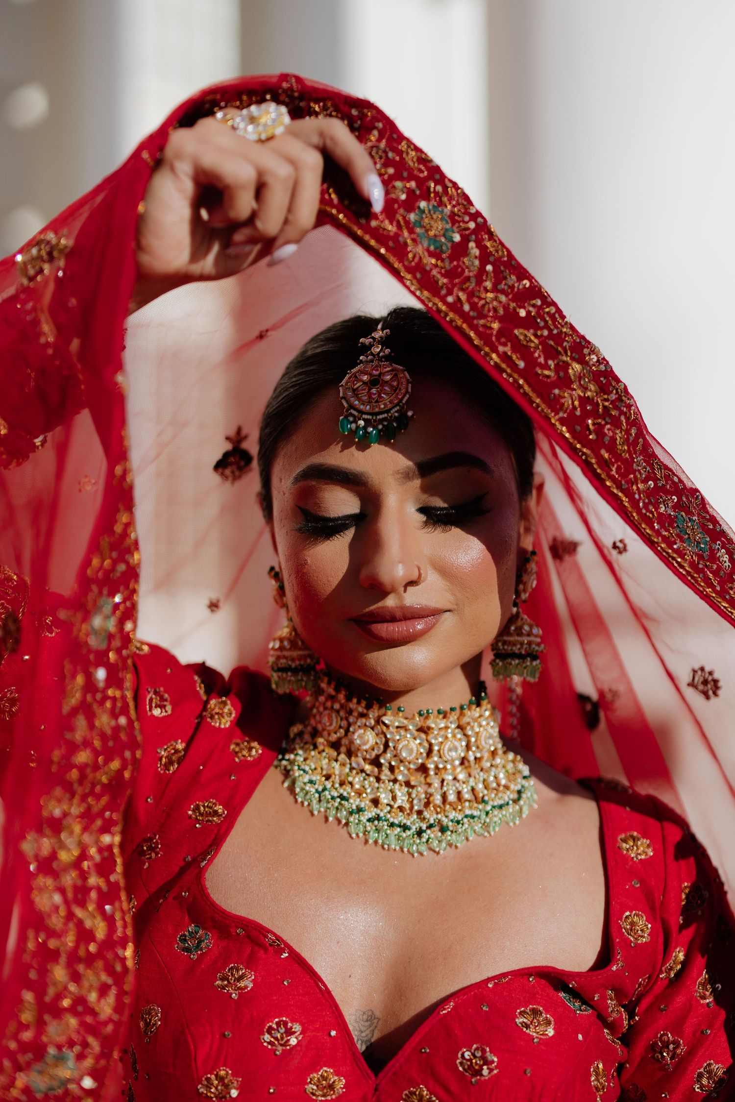 A bride in traditional South Asian wedding attire wearing a red embroidered dress with gold jewelry and a flowing veil.