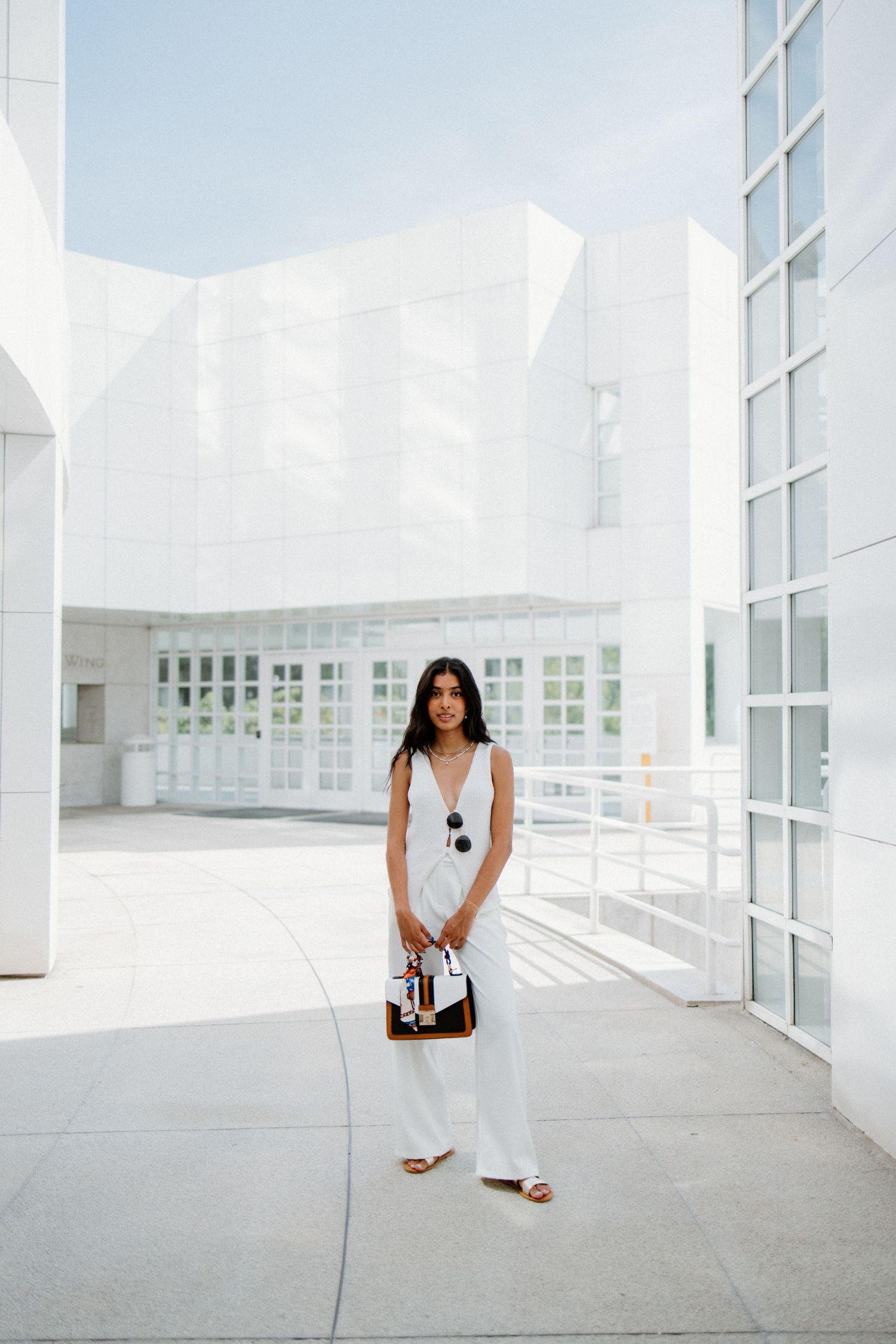 A figure in a sleek white outfit stands in a modern minimalist architectural space with bright white walls and glass windows.